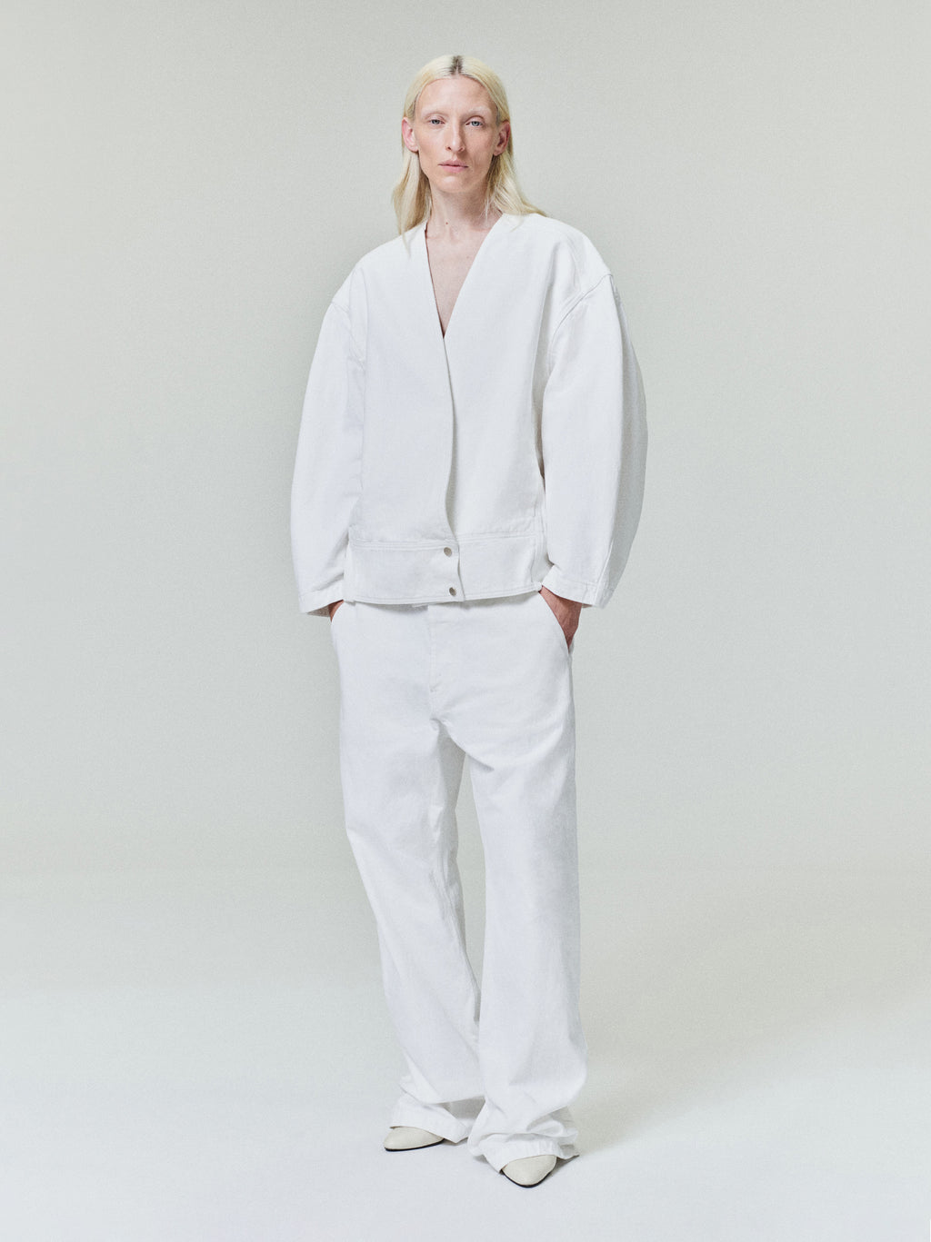 Person with long light blonde hair wearing loose white jacket with wide white pants and white shoes on pale background