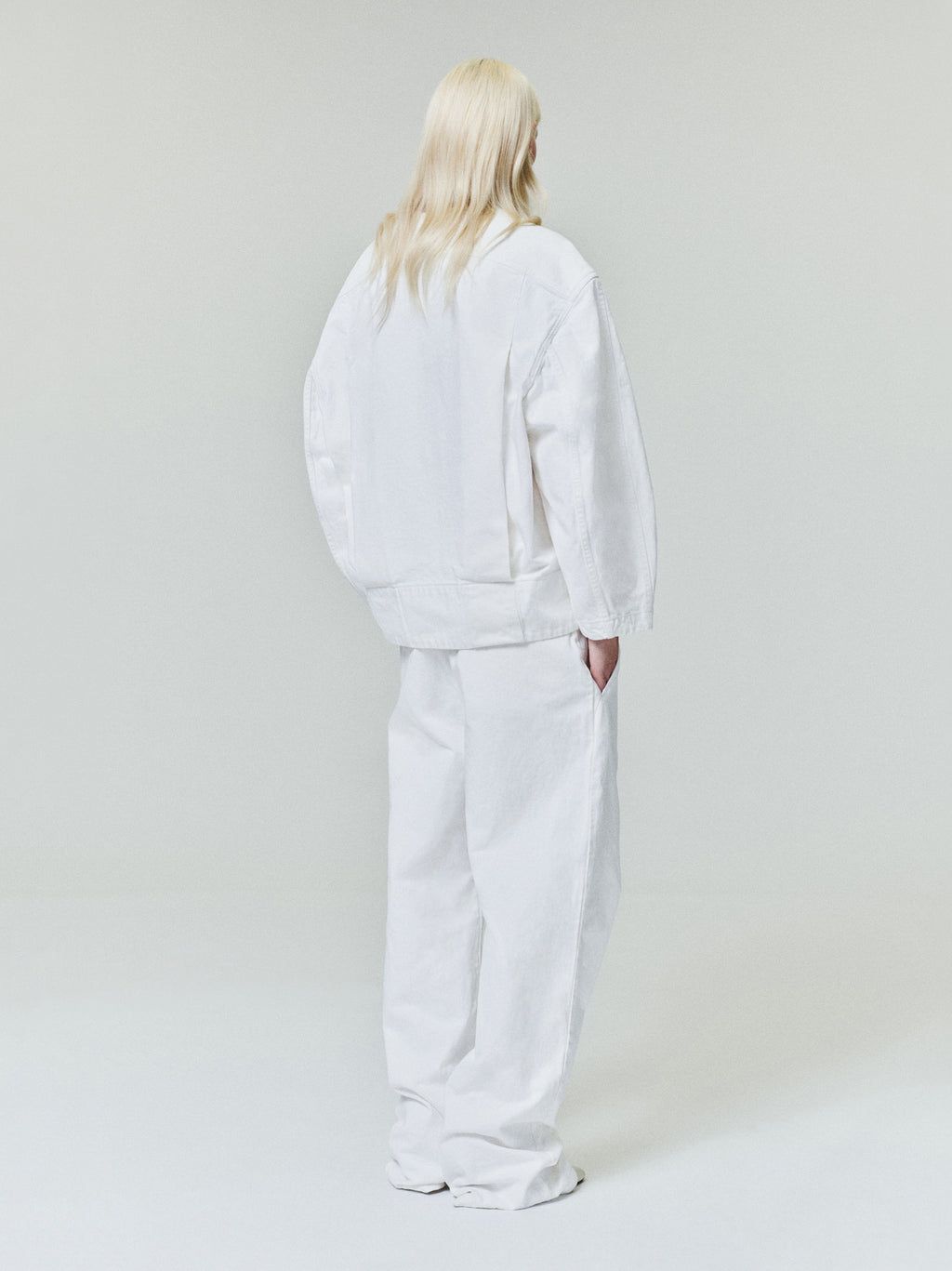 Person with long light blonde hair wearing oversized white jacket and wide white pants standing against plain light background
