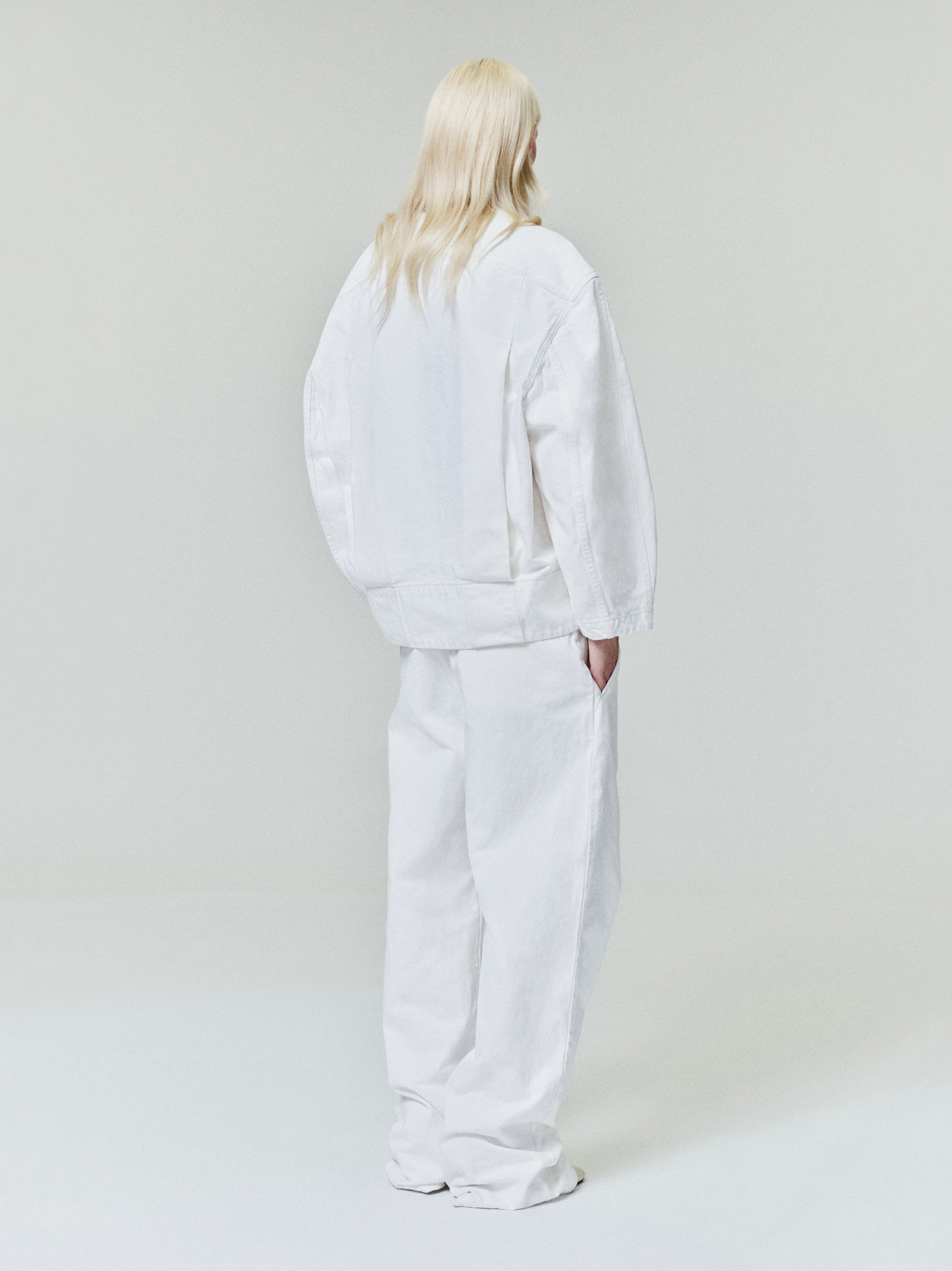 Person with long light blonde hair wearing oversized white jacket and wide white pants standing against plain light background