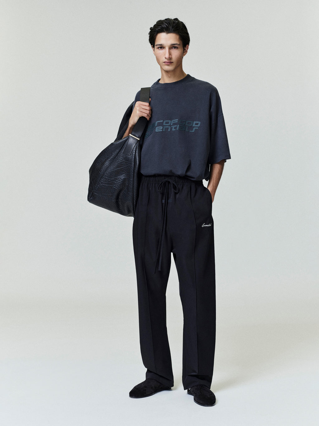 Person wearing a dark outfit with a modern style Fear Of God Essentials logo, holding a black bag on a plain background