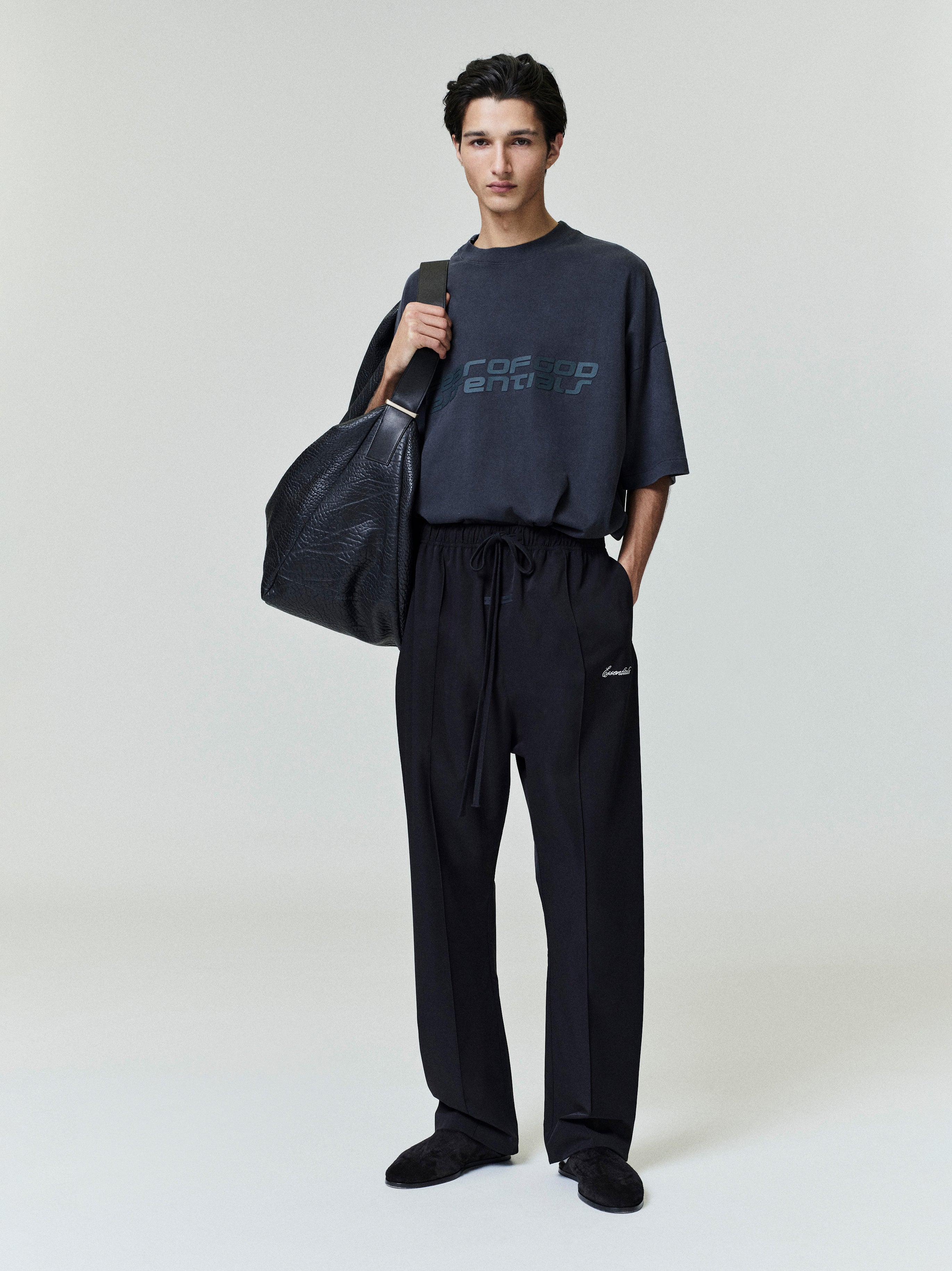 Person wearing a dark outfit with a modern style Fear Of God Essentials logo, holding a black bag on a plain background