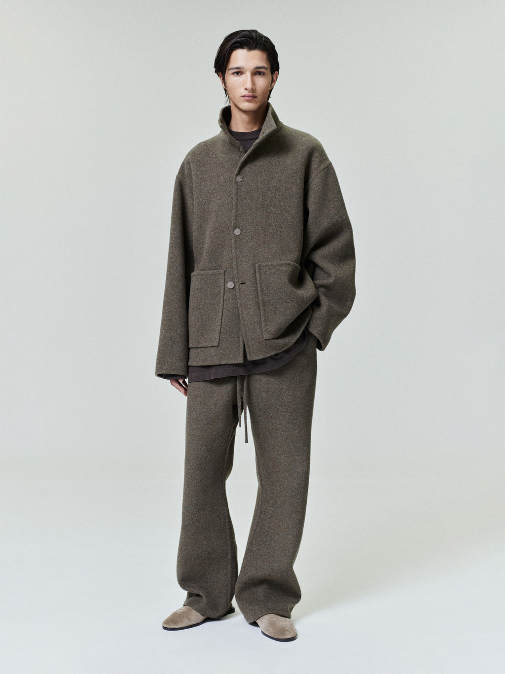 Man wearing a loose brown button-up jacket with large front pockets and matching wide-legged pants standing against a plain backdrop