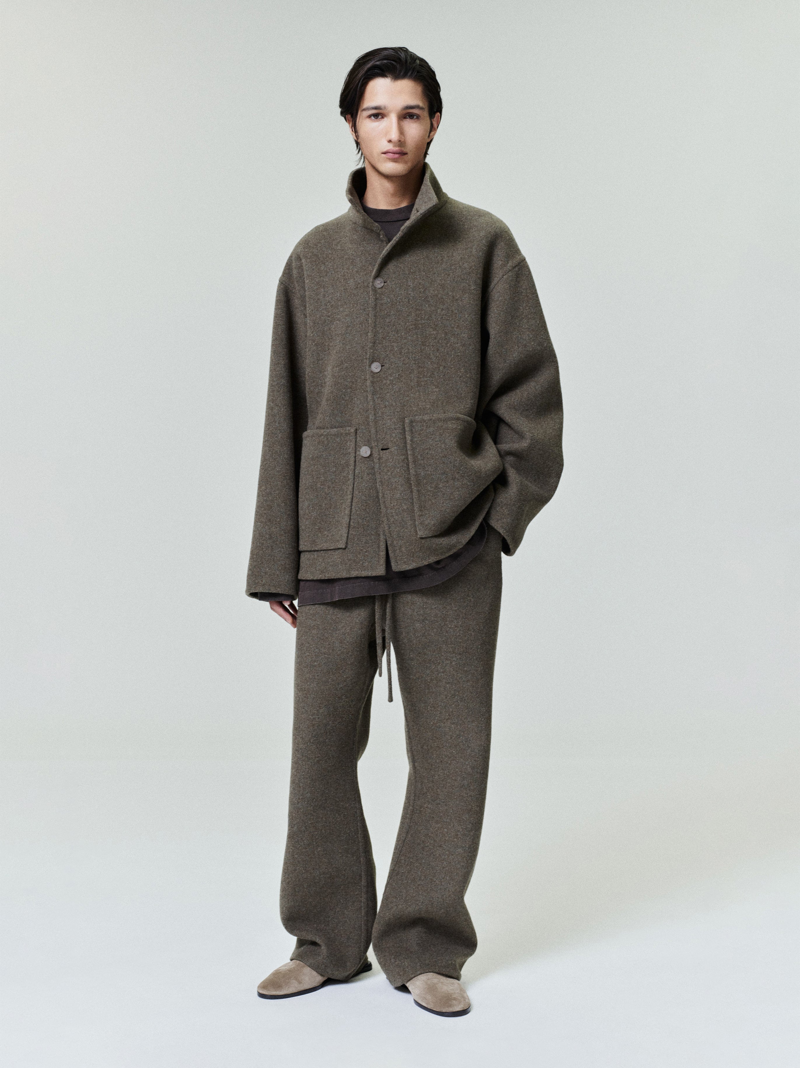 Man wearing a loose brown button-up jacket with large front pockets and matching wide-legged pants standing against a plain backdrop
