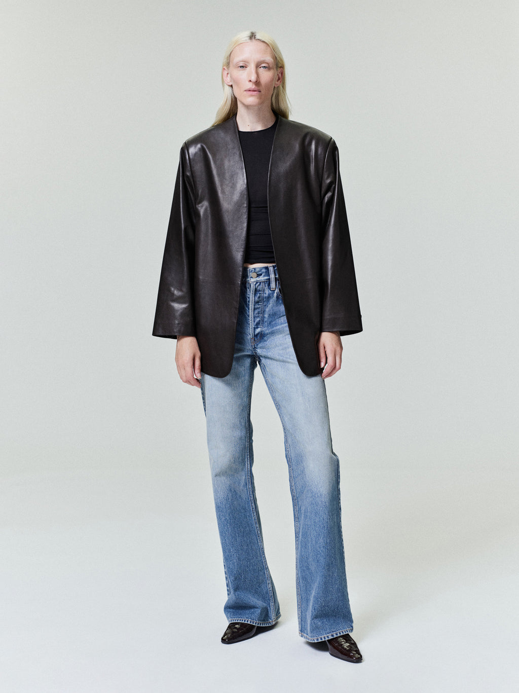 Person with straight pale blonde hair wearing oversized black leather jacket and faded blue flared jeans on light gray background