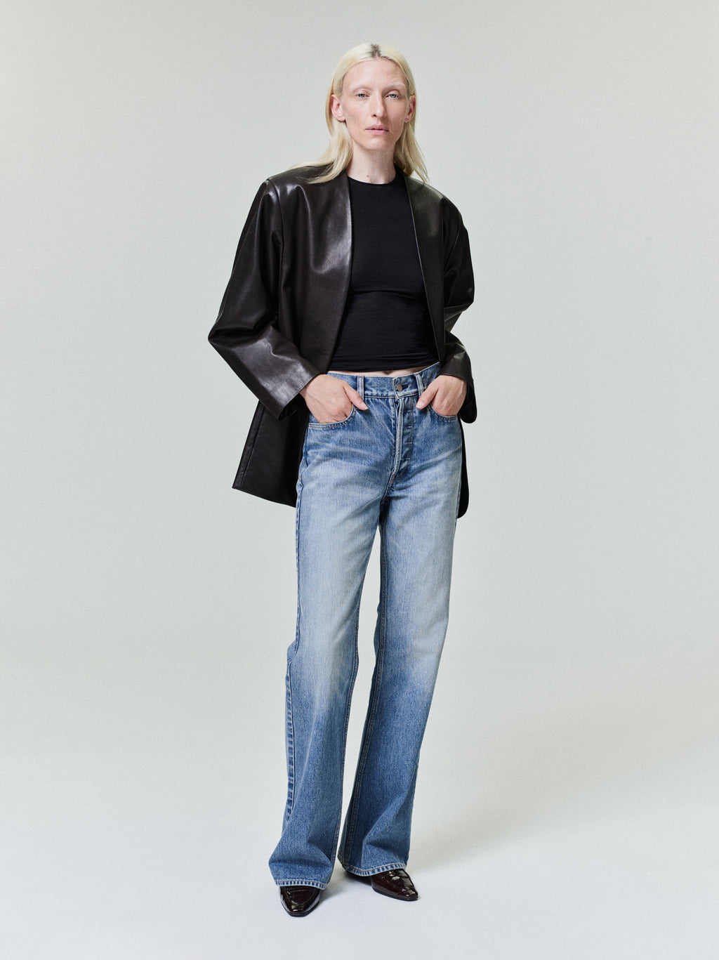 Person with straight light blonde hair wearing black cropped top, black leather jacket, and faded blue jeans on pale background