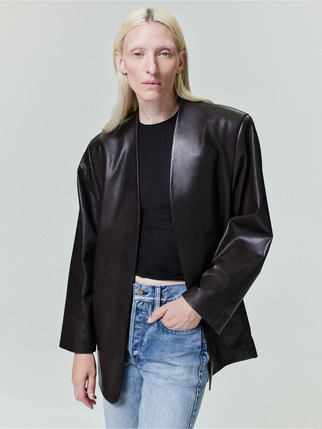 Person with straight pale blonde hair wearing oversized black leather jacket and light blue jeans on plain background