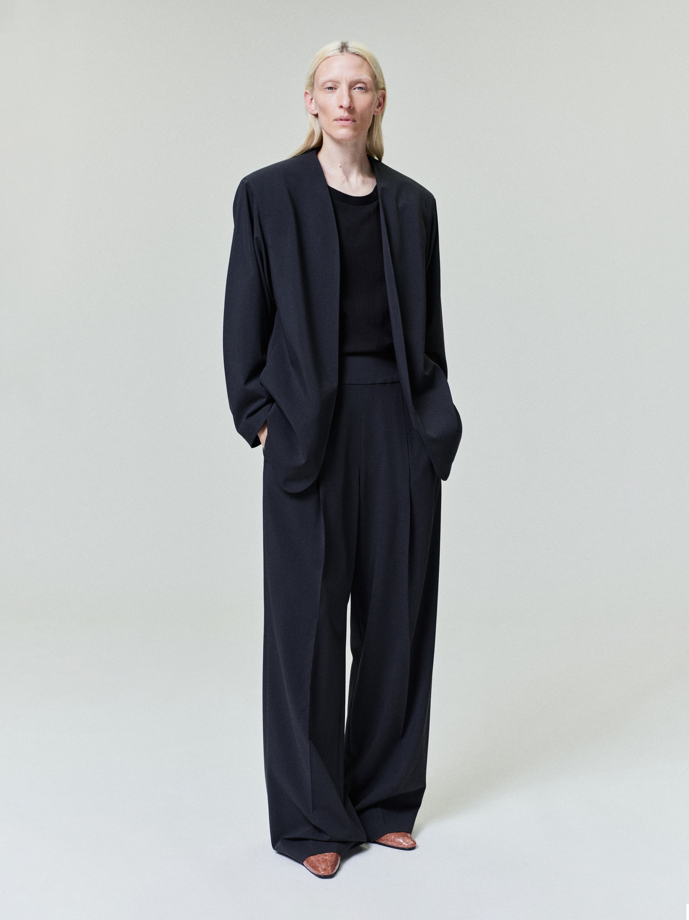 Person with pale blonde hair wearing loose-fitting black blazer and wide-leg black trousers on light gray background