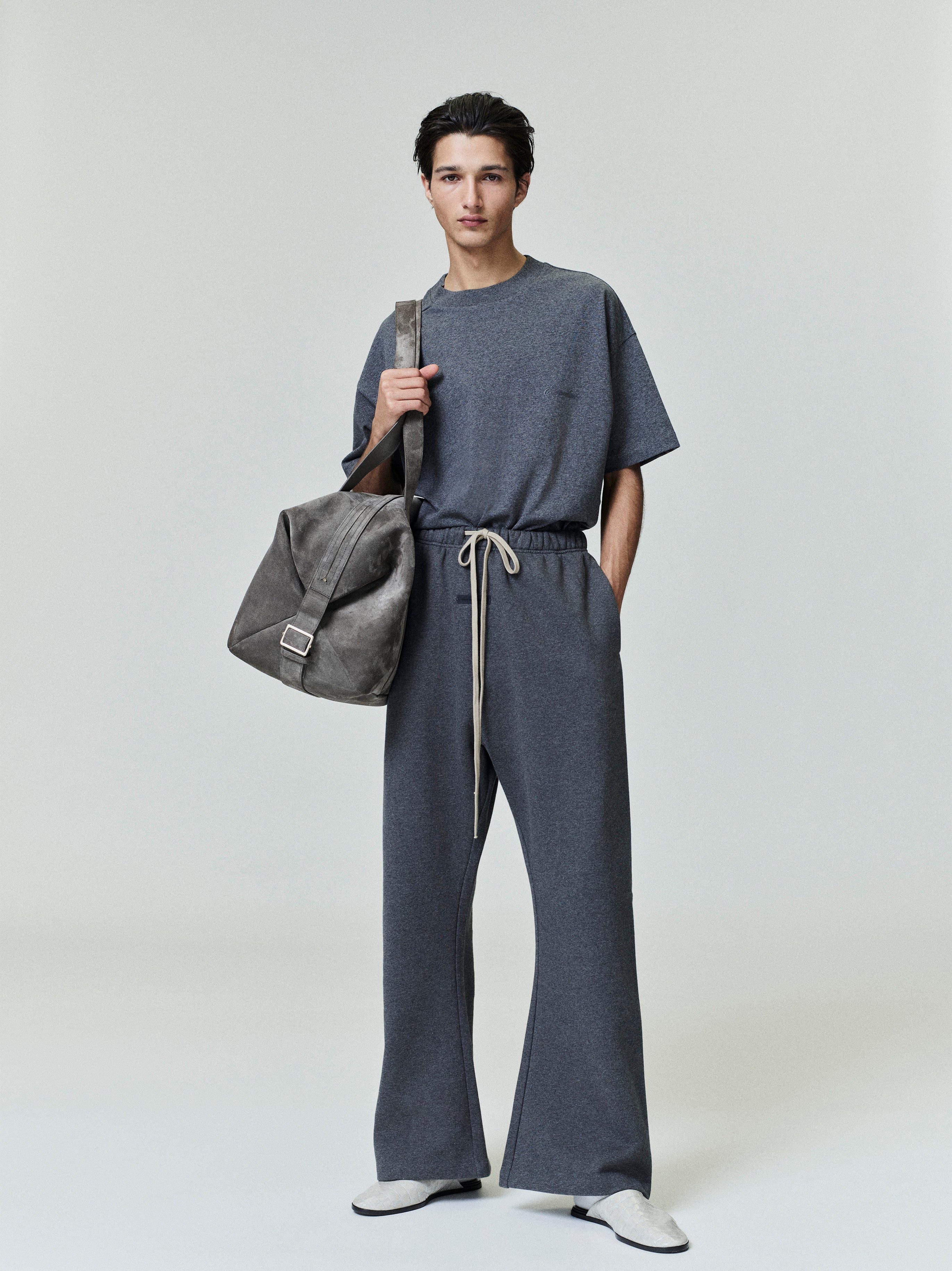 Model wearing dark grey flare sweatpants with beige drawstrings, paired with matching short-sleeve top and carrying gray bag front view