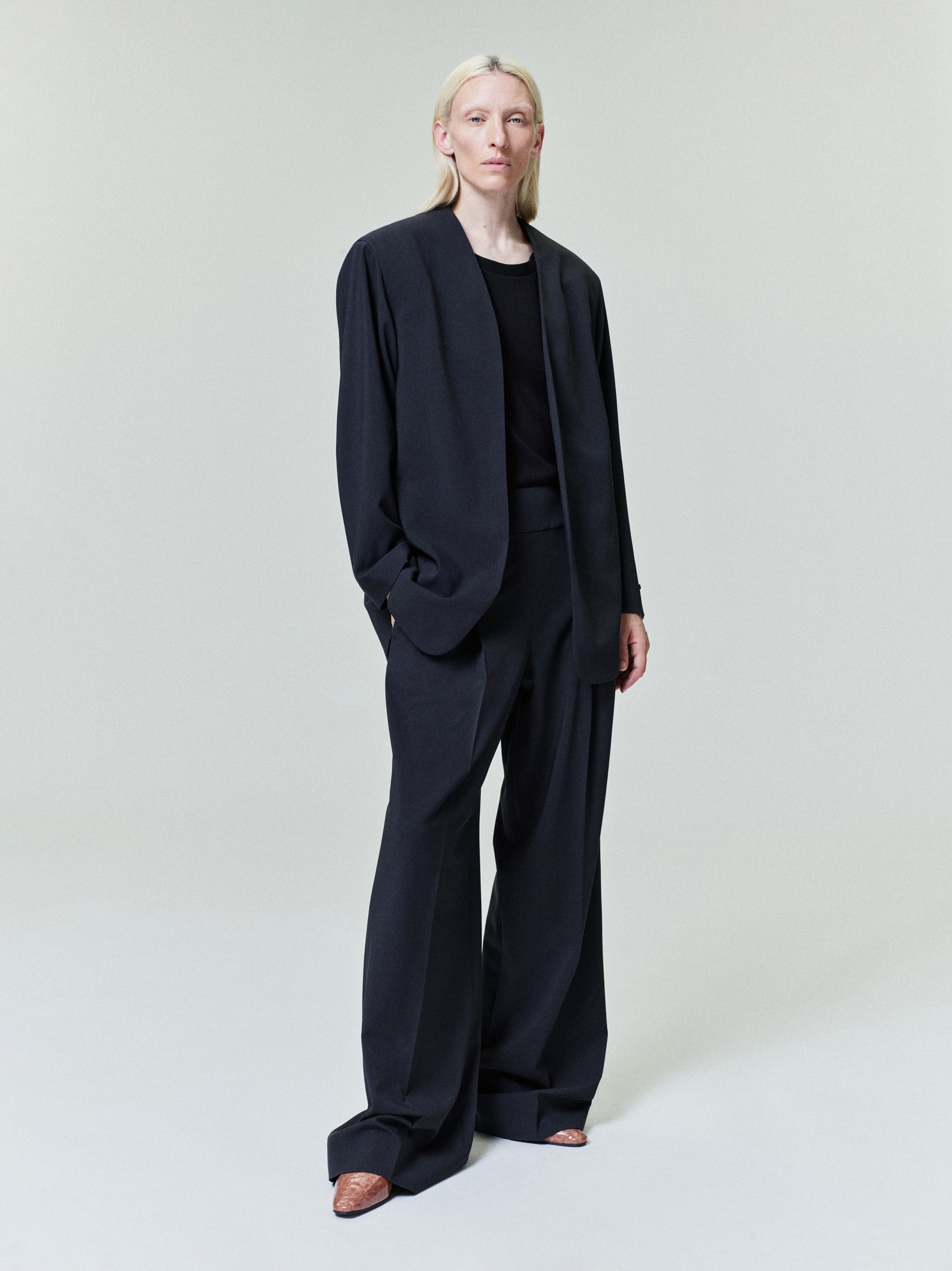 Person with pale blonde hair wearing relaxed black blazer and wide-leg black trousers with brown shoes on light background