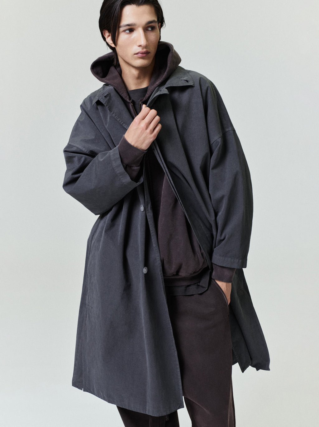 Young man wearing a loose dark gray trench coat layered over a dark hoodie and matching pants against a plain backdrop