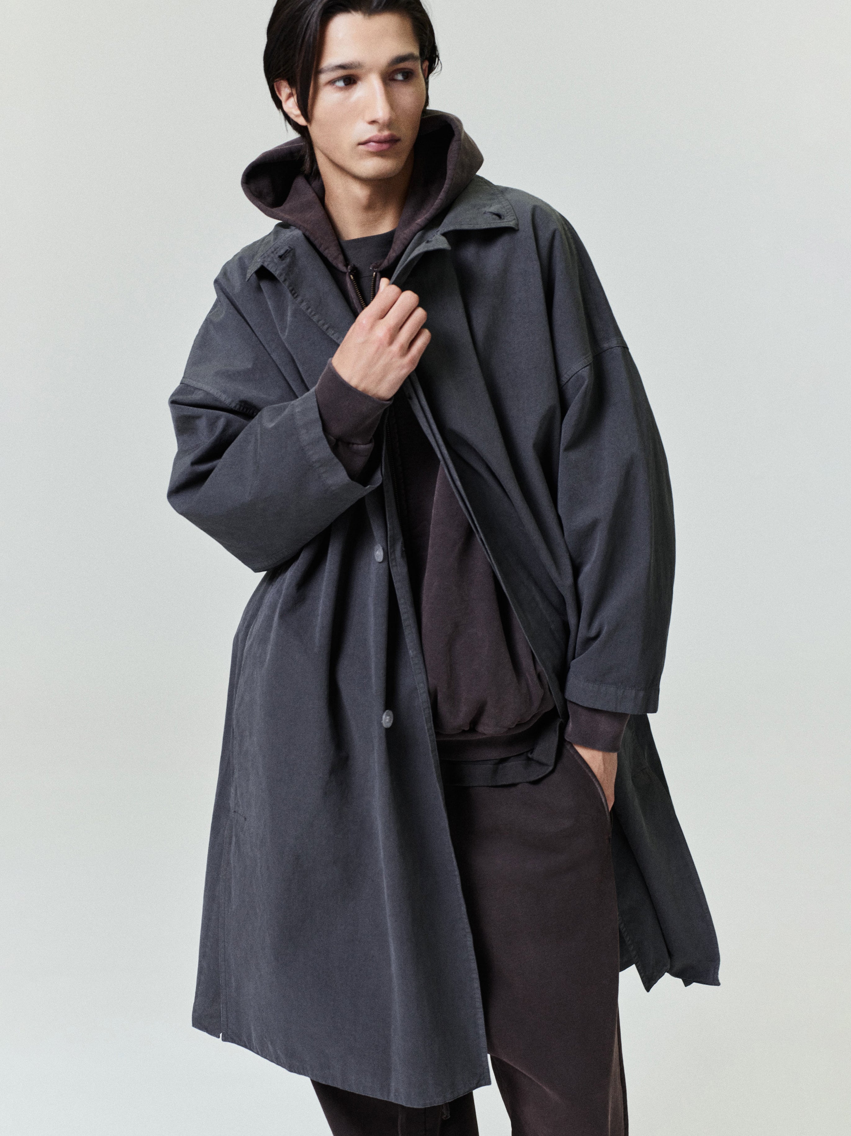 Young man wearing a loose dark gray trench coat layered over a dark hoodie and matching pants against a plain backdrop
