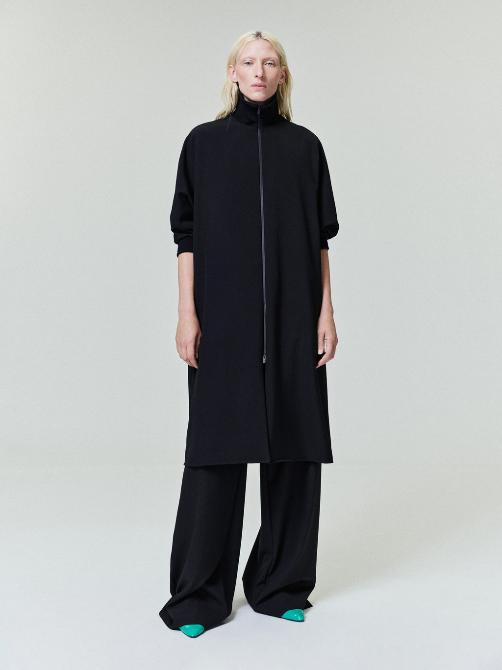 Woman wearing a long black zip-up coat with elbow-length sleeves and wide-leg black pants against a plain backdrop.