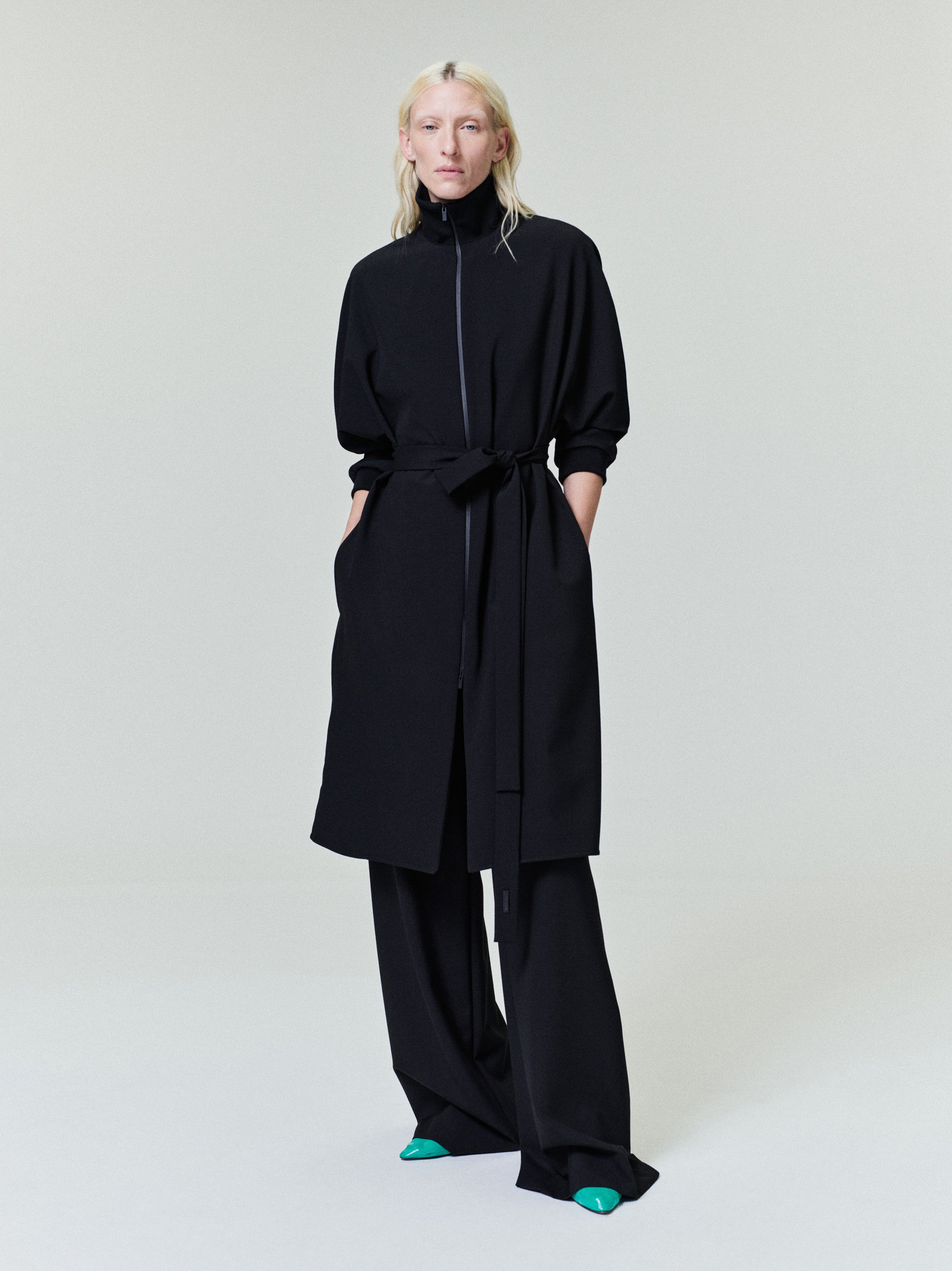 Model wearing black long coat with tied waist belt and wide-leg black pants against a plain light background.