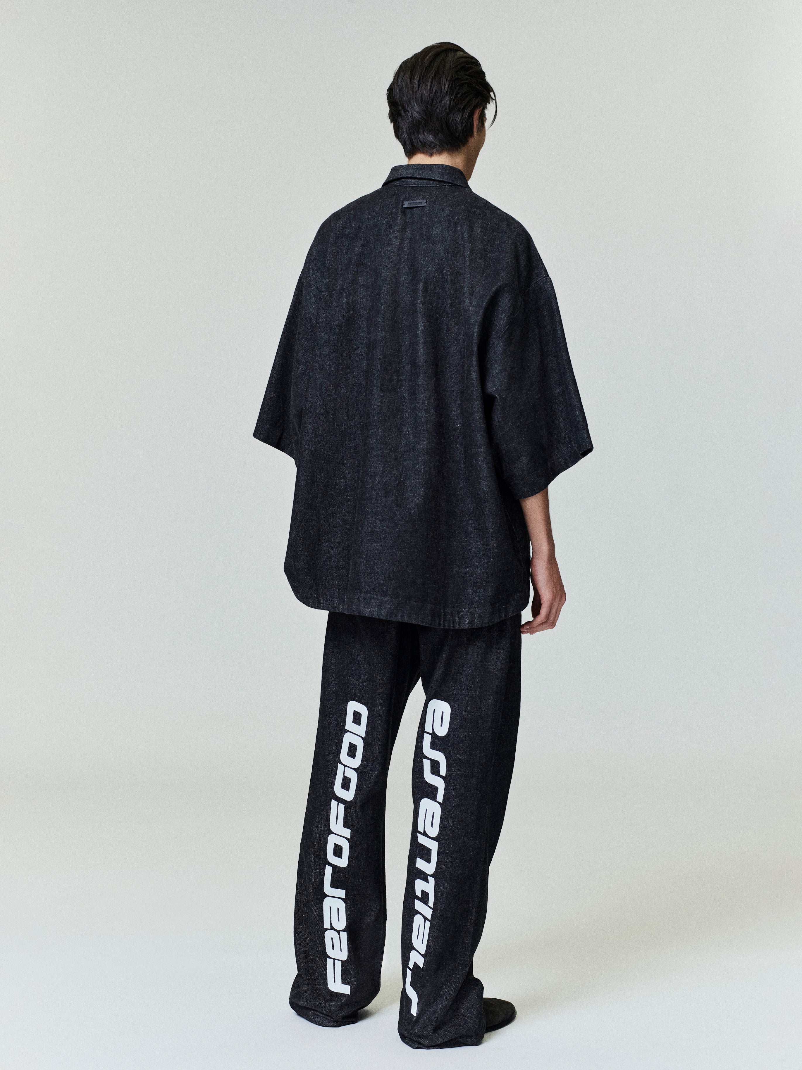 Rear view of model wearing black denim work pants with large white vertical text and oversized dark shirt