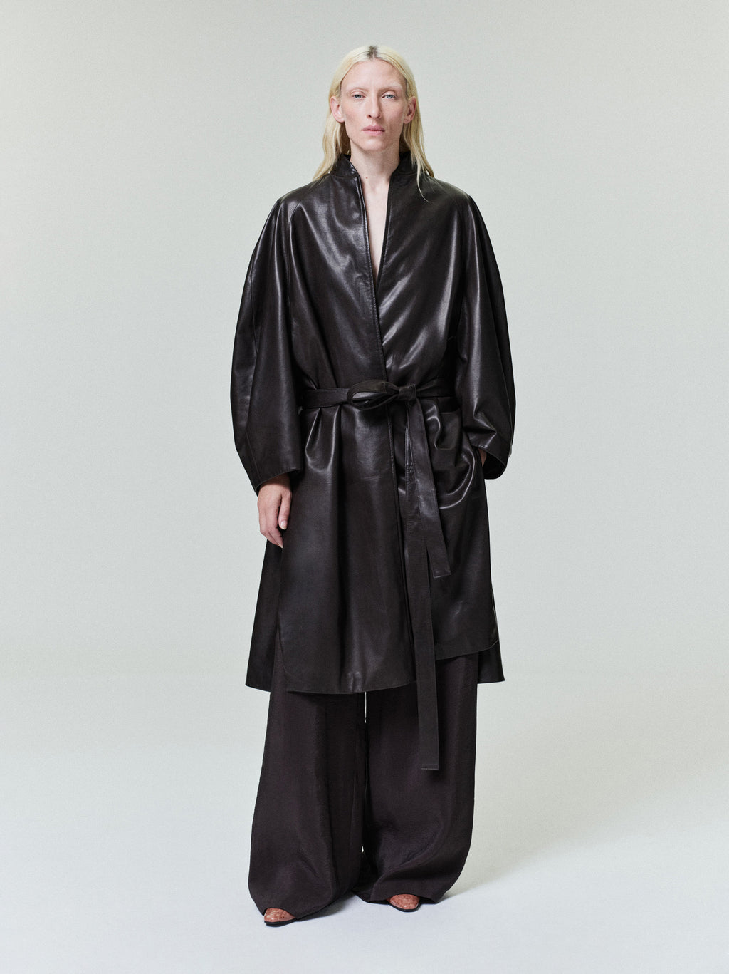 Person with long pale blonde hair wearing a belted black leather coat and wide black pants on plain background