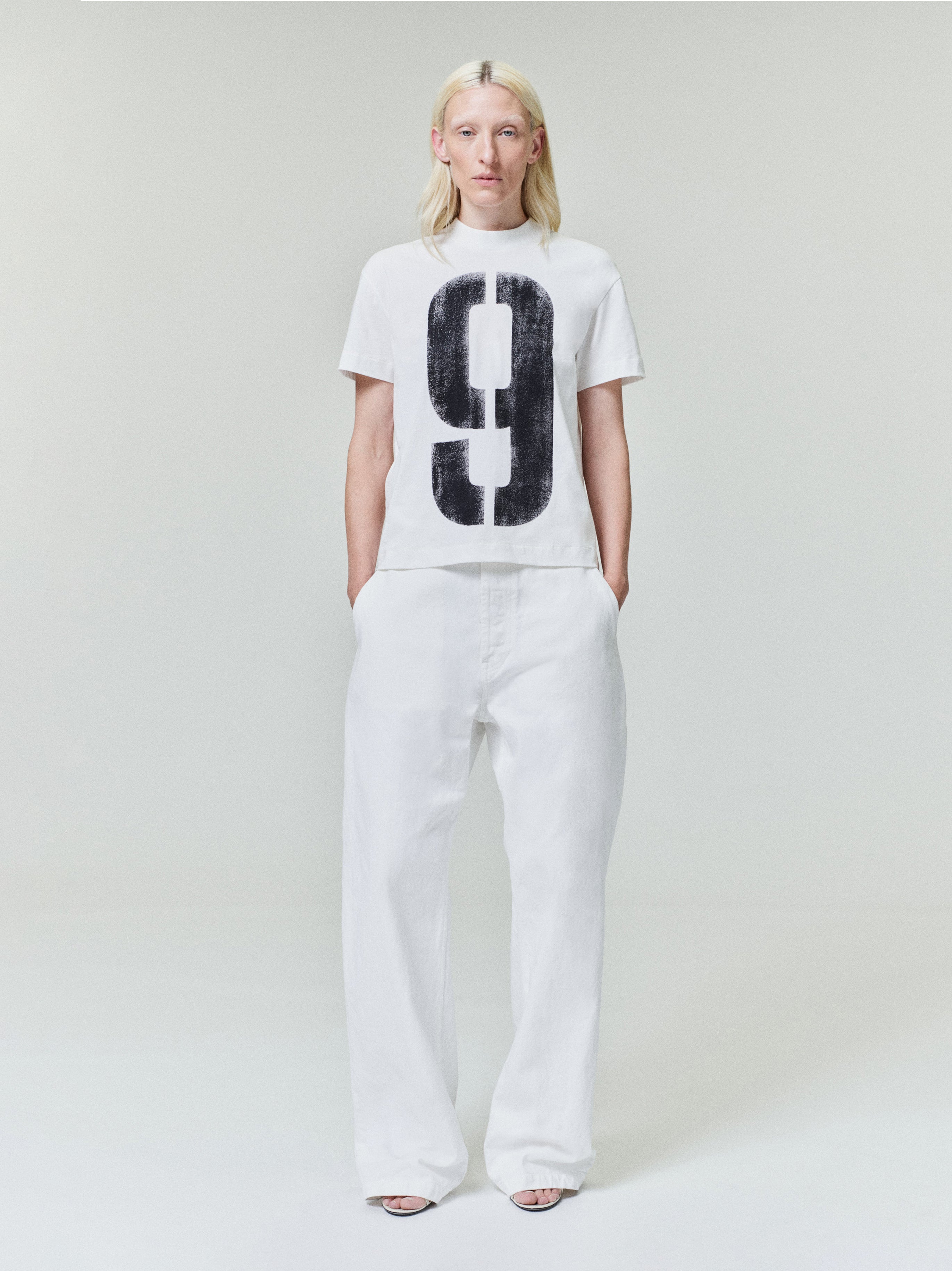 Person with straight blonde hair wearing white t-shirt featuring large black number 9 and loose white wide-leg pants on light background