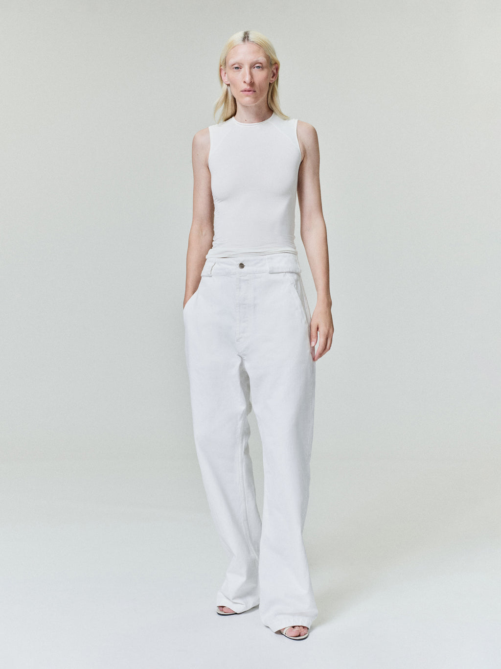 Person with pale blonde hair wearing sleeveless white top and high-waisted loose white pants on plain light background
