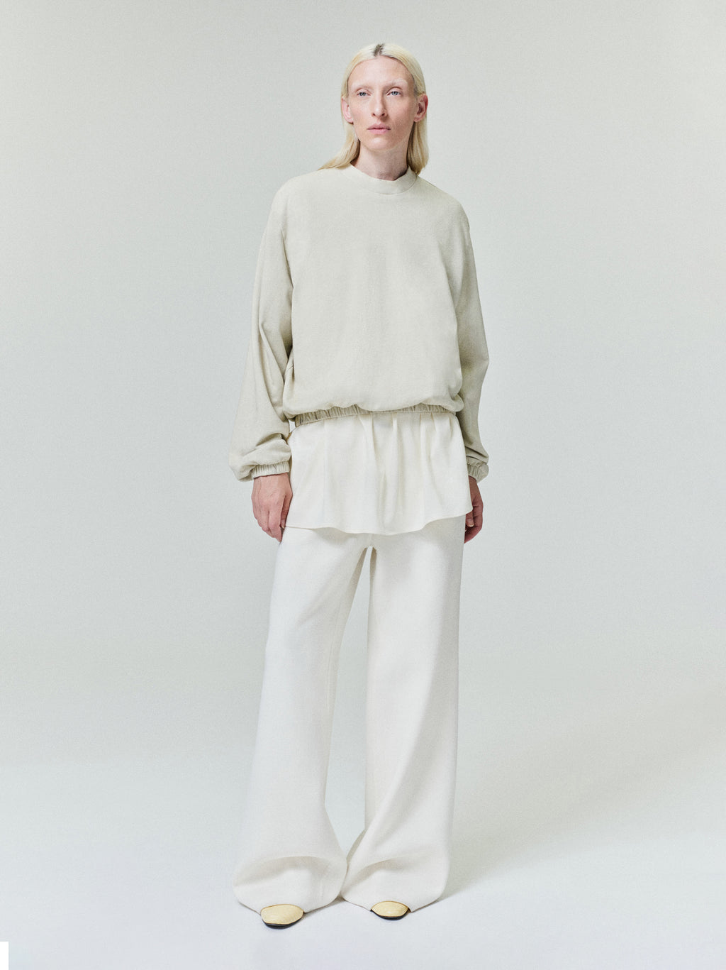 Person with blonde hair wearing light beige loose long-sleeve top layered over white wide-leg pants on pale background