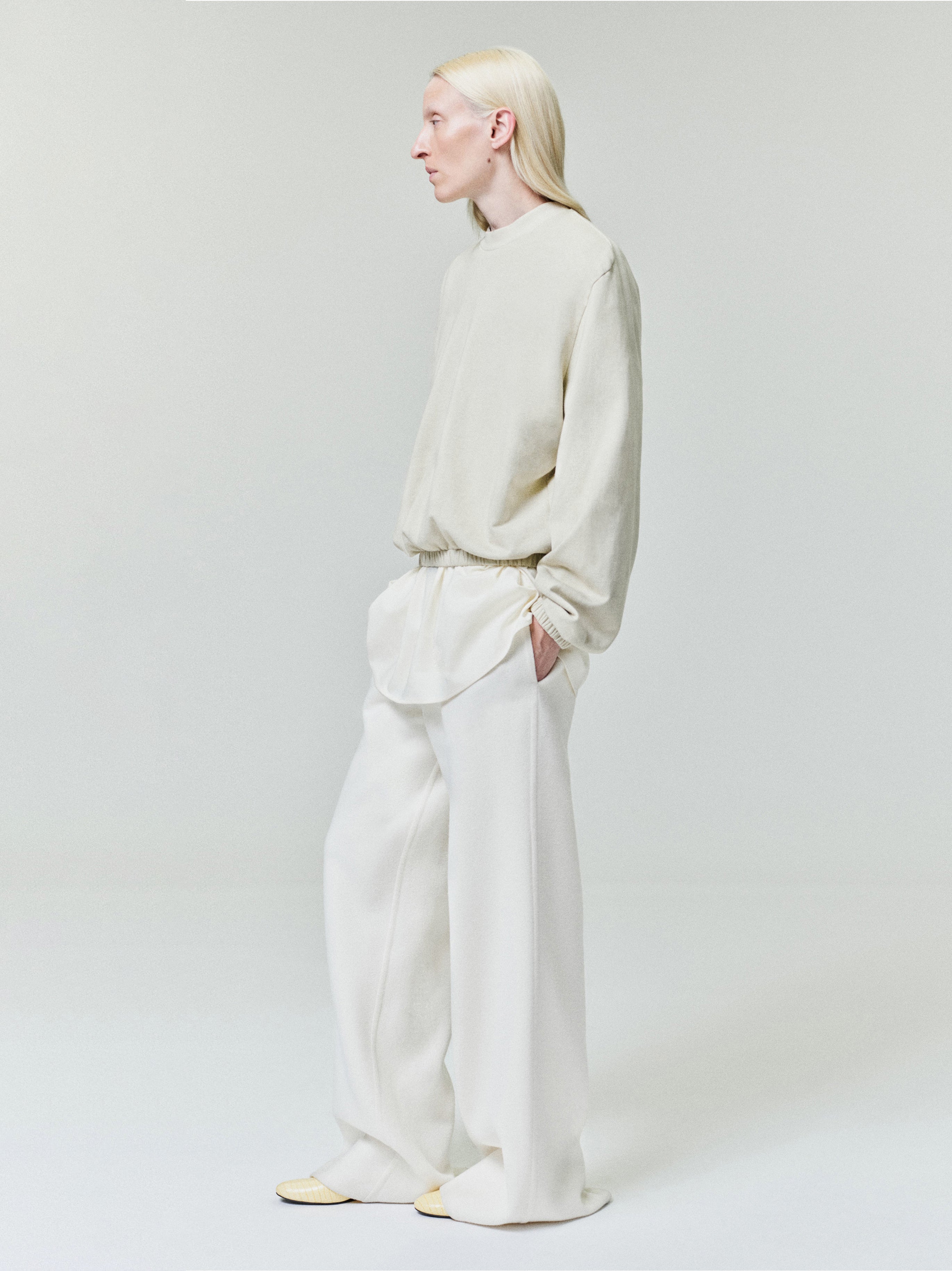 Person with long blonde hair wearing light beige loose sweater over white shirt and wide white pants on pale background
