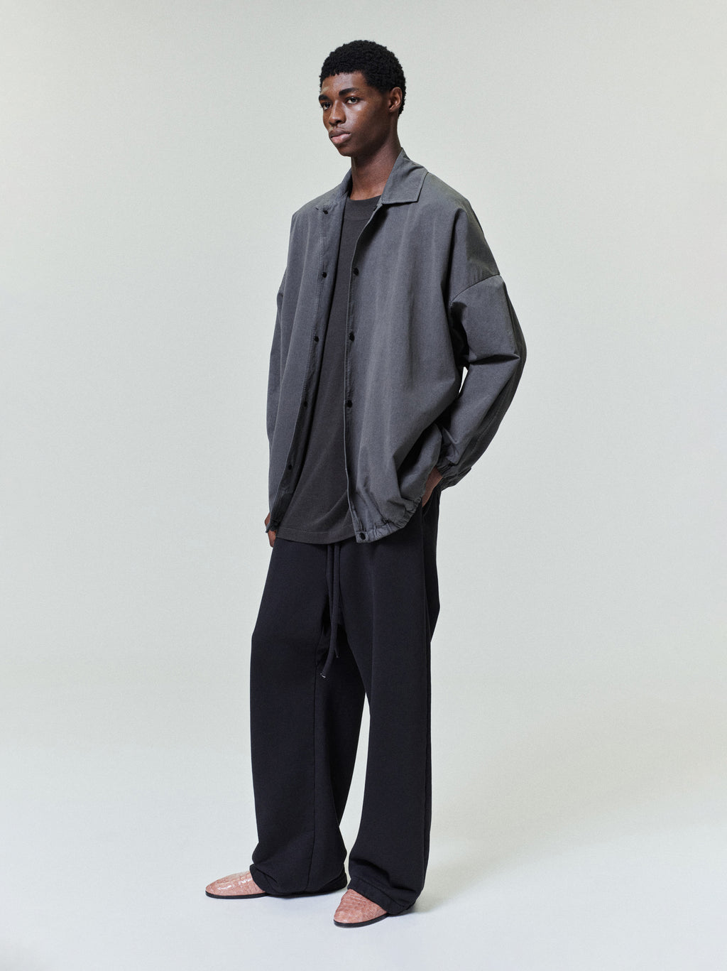 Young man wearing a loose gray button-up jacket over a black shirt with wide black trousers on a neutral background
