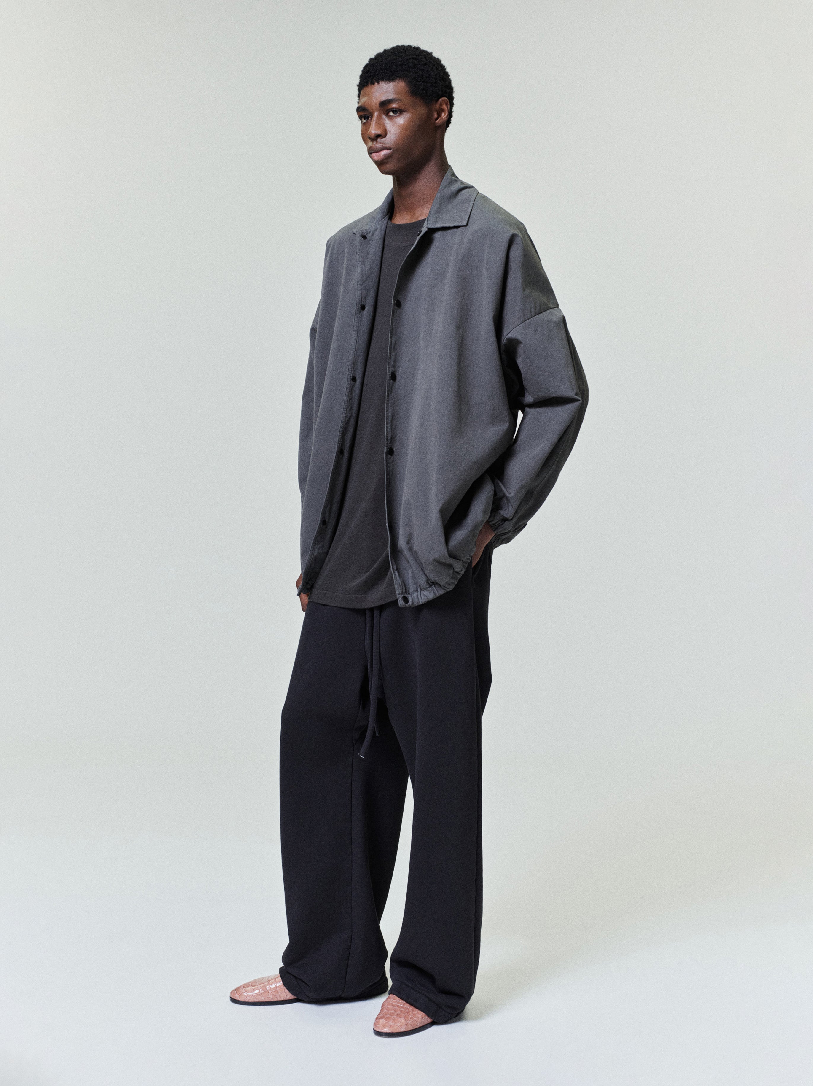 Young man wearing a loose gray button-up jacket over a black shirt with wide black trousers on a neutral background