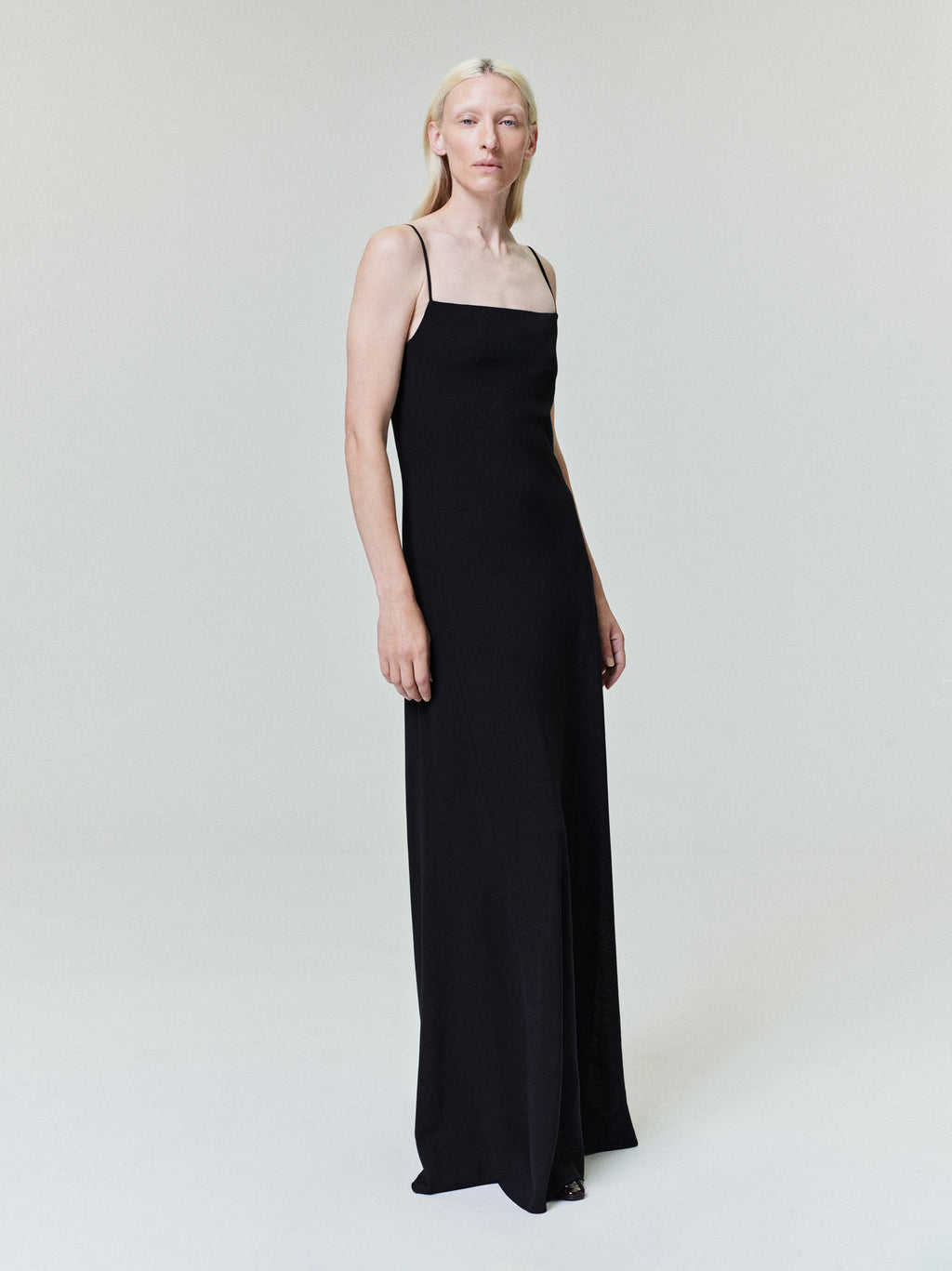 Person with pale blonde hair wearing long black sleeveless dress with thin straps on plain light background