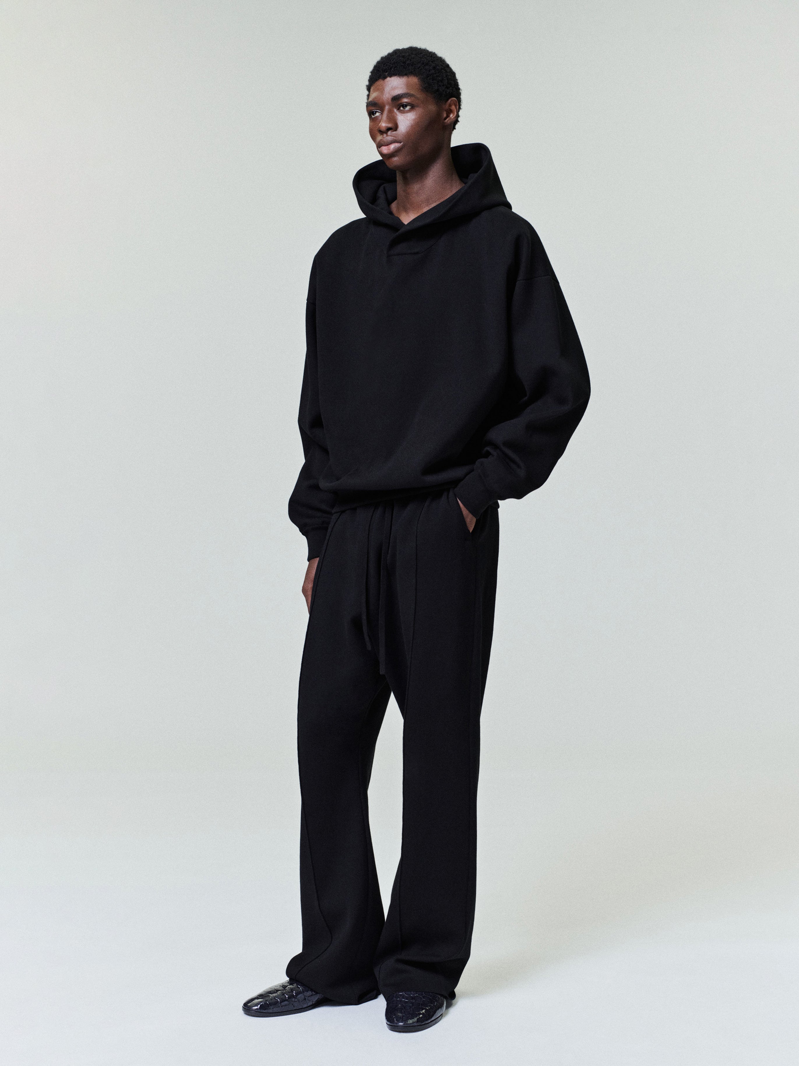 Male model wearing loose black hoodie and matching wide-leg pants with hands in pockets on plain background