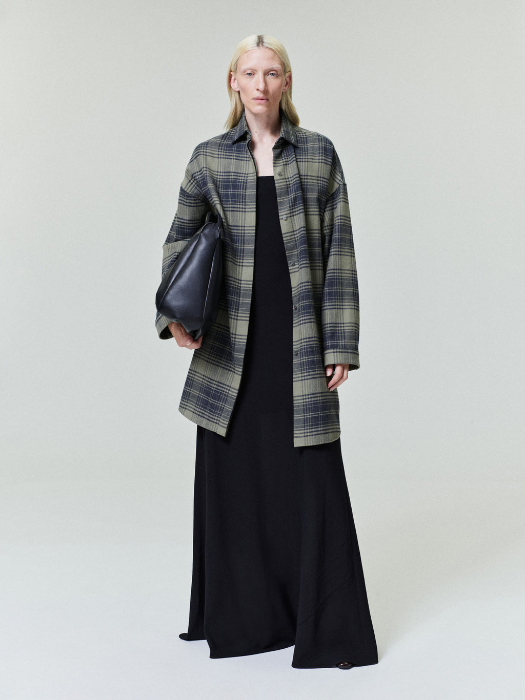 Model wearing an oversized green and black plaid shirt jacket over a long black dress holding a black leather bag