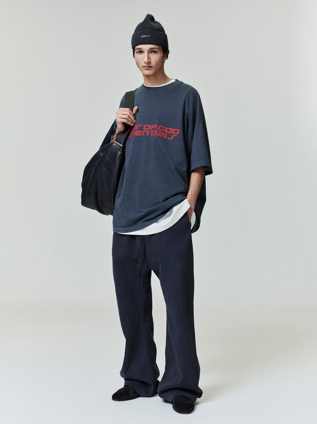 Full body view of model wearing navy Fear of God Essentials 90s Tee with red logo and relaxed fit, styled with black beanie and bag