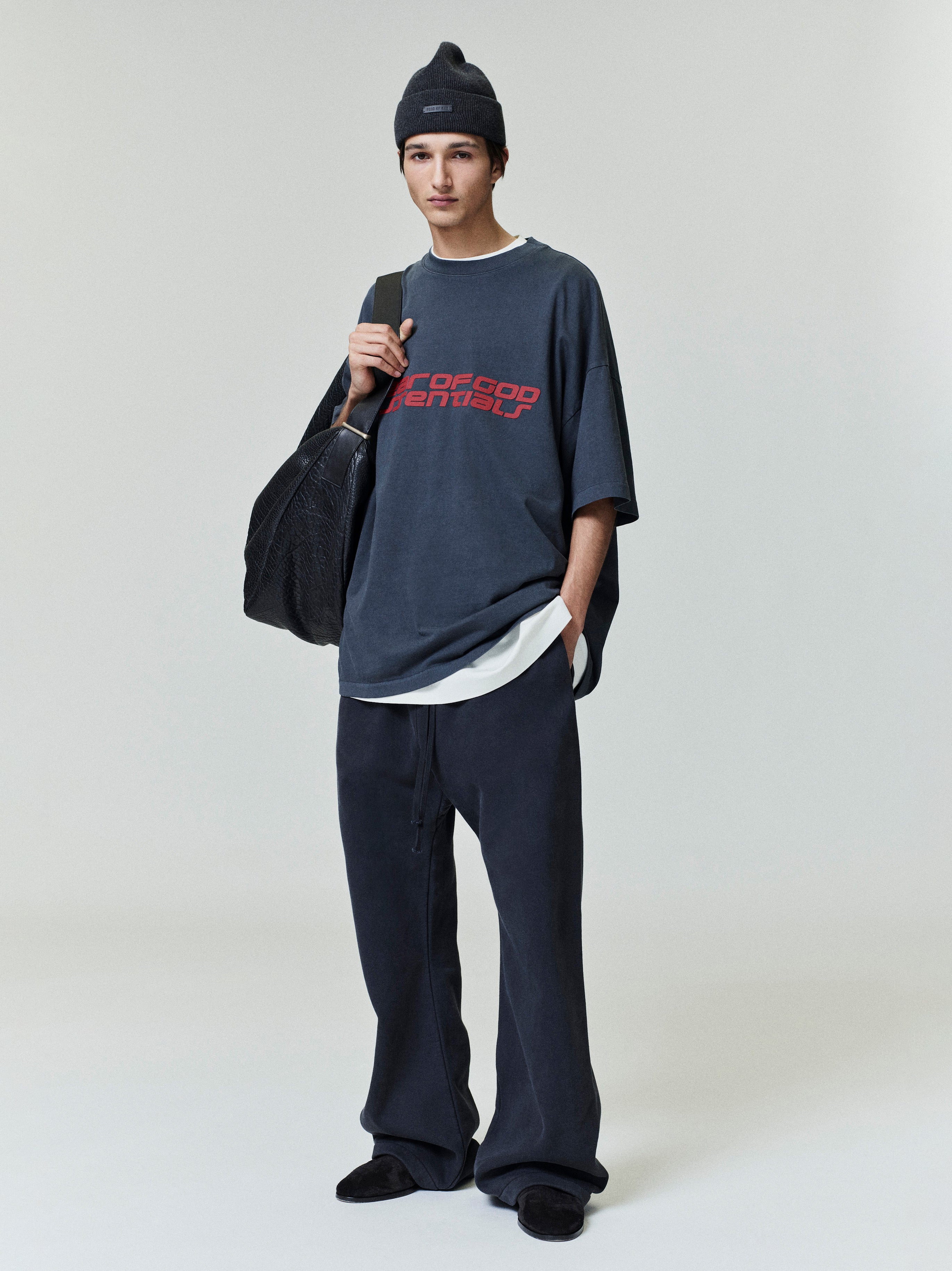Full body view of model wearing navy Fear of God Essentials 90s Tee with red logo and relaxed fit, styled with black beanie and bag