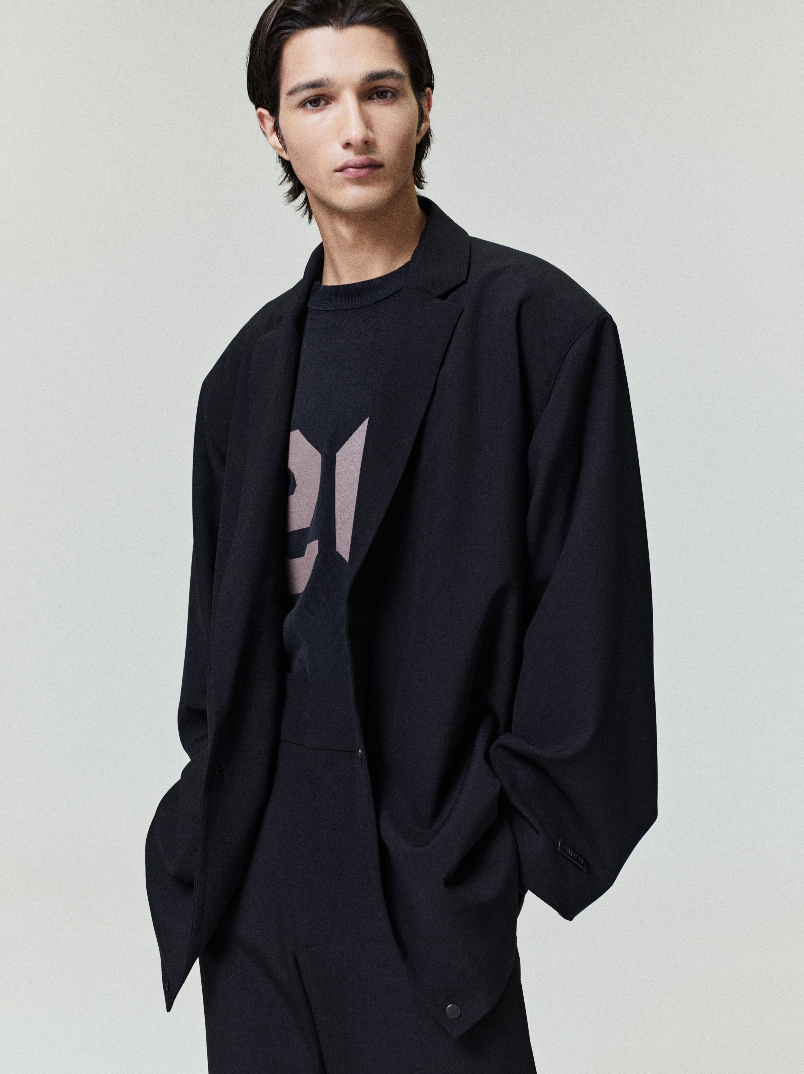 Young man wearing a loose black blazer over a black graphic t-shirt and matching black trousers with hands in pockets