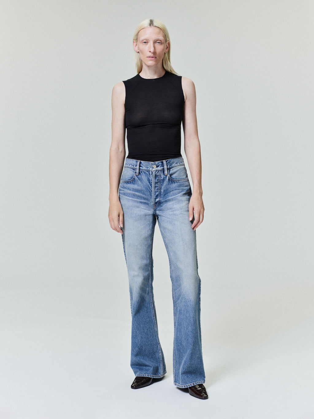 Person with straight pale blonde hair wearing fitted sleeveless black top and light blue flared jeans on plain background