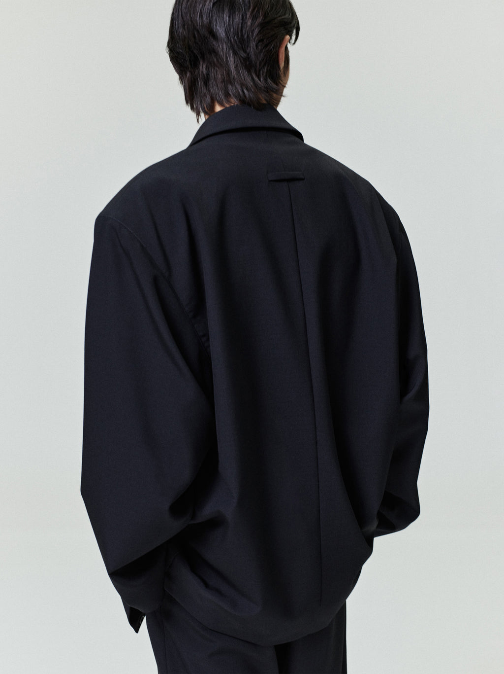 Man seen from behind wearing an oversized black jacket with dropped shoulders and a back pleat detail