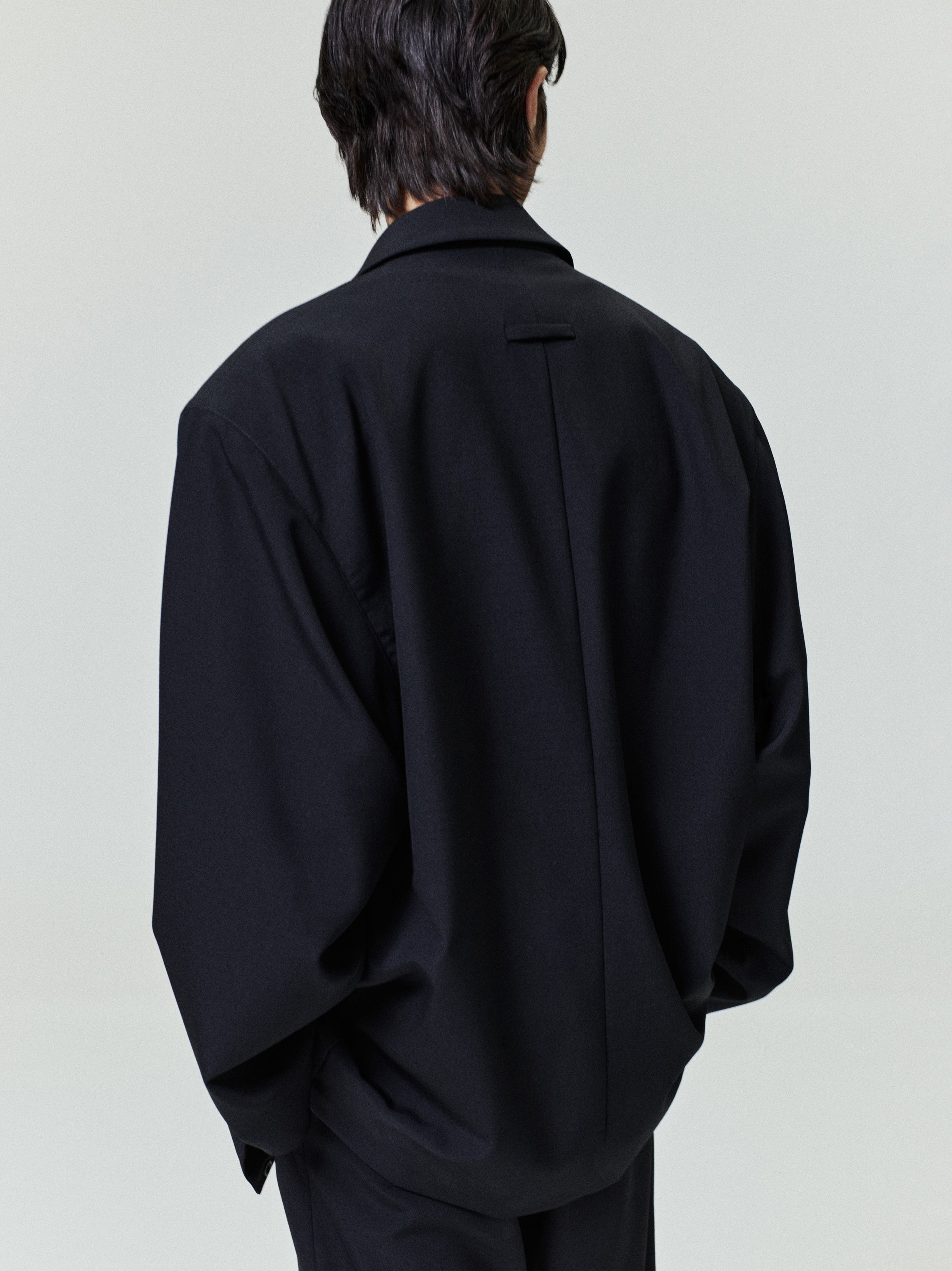 Man seen from behind wearing an oversized black jacket with dropped shoulders and a back pleat detail
