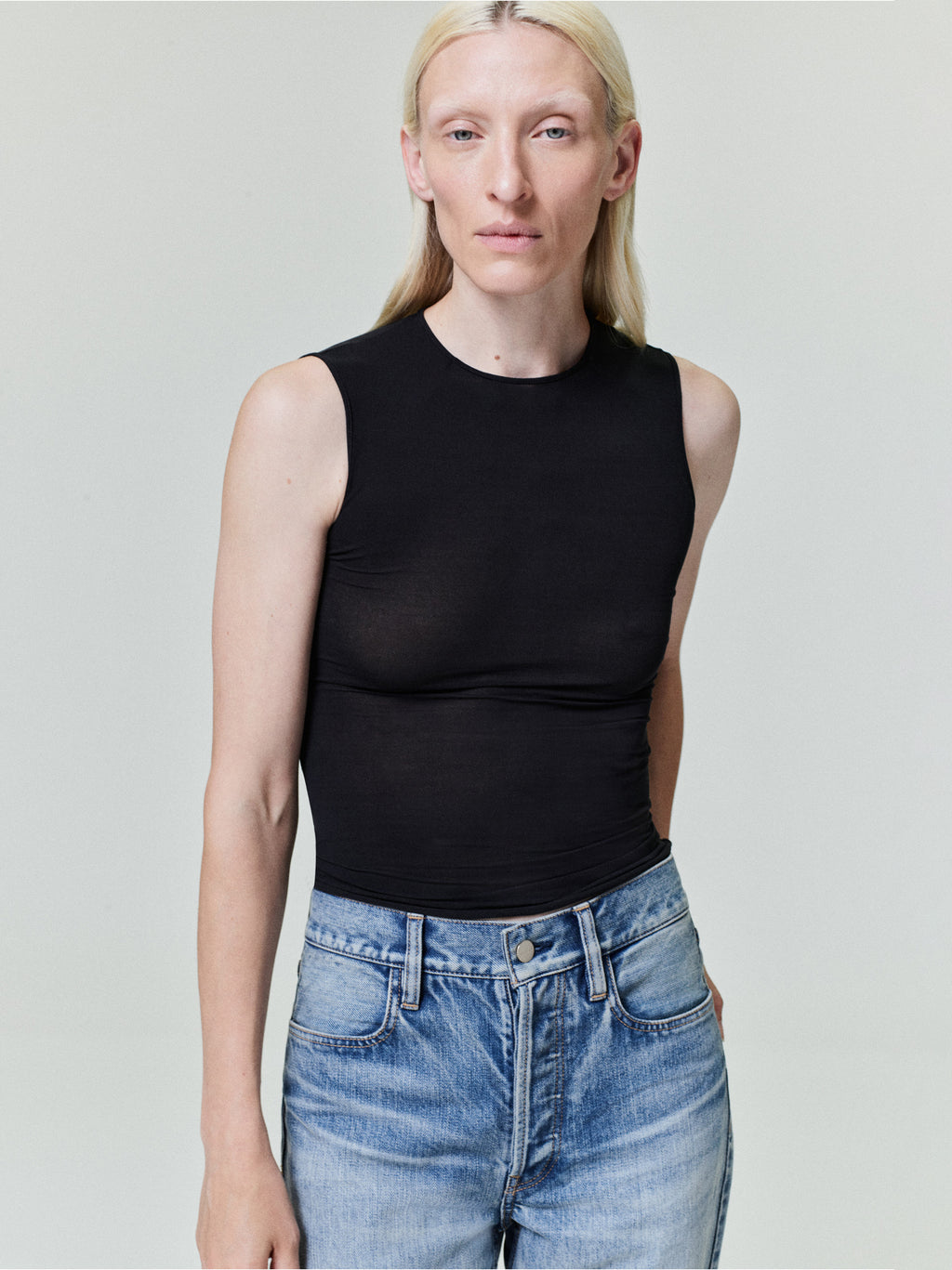 Person with straight pale blonde hair wearing fitted sleeveless black top and high-waisted light blue jeans on plain background