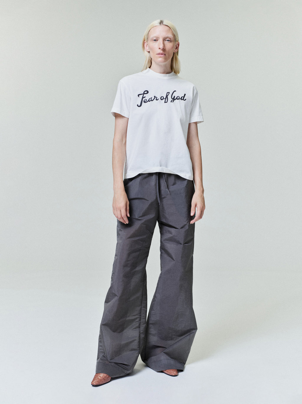 Person with straight blonde hair wearing white "Fear of God" t-shirt and loose dark gray wide-leg pants standing on light background