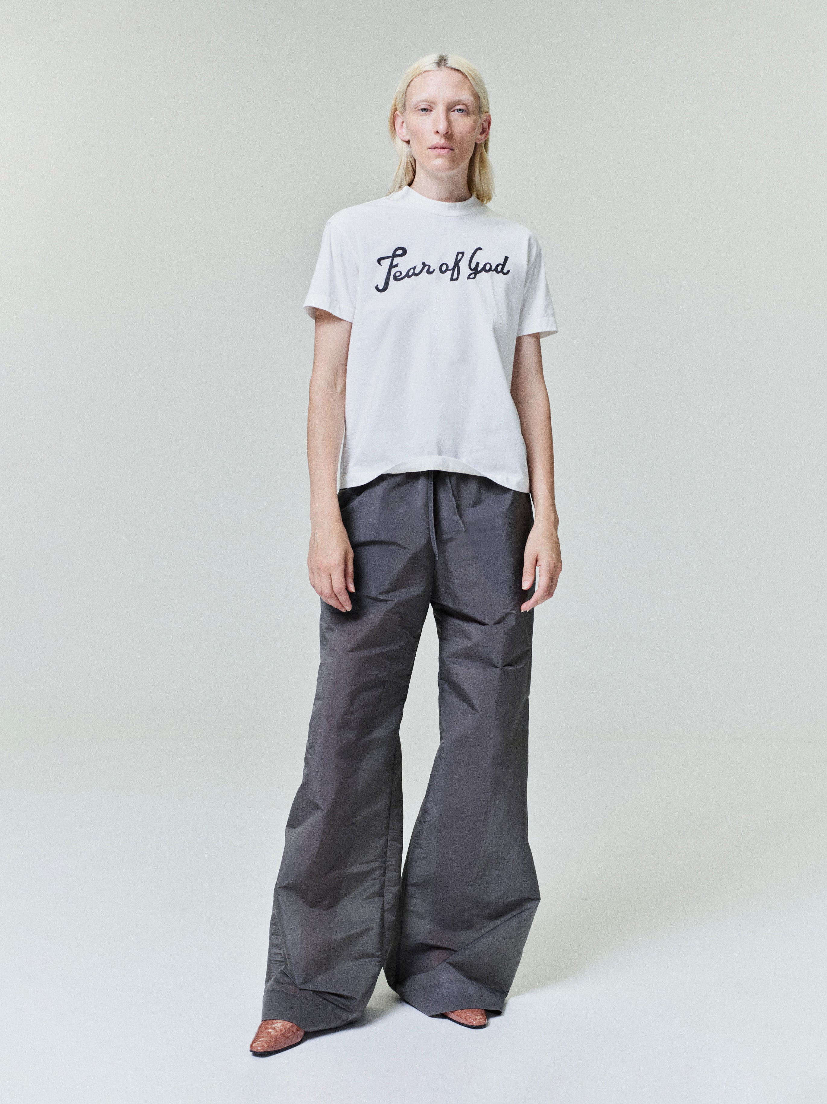 Person with straight blonde hair wearing white "Fear of God" t-shirt and loose dark gray wide-leg pants standing on light background