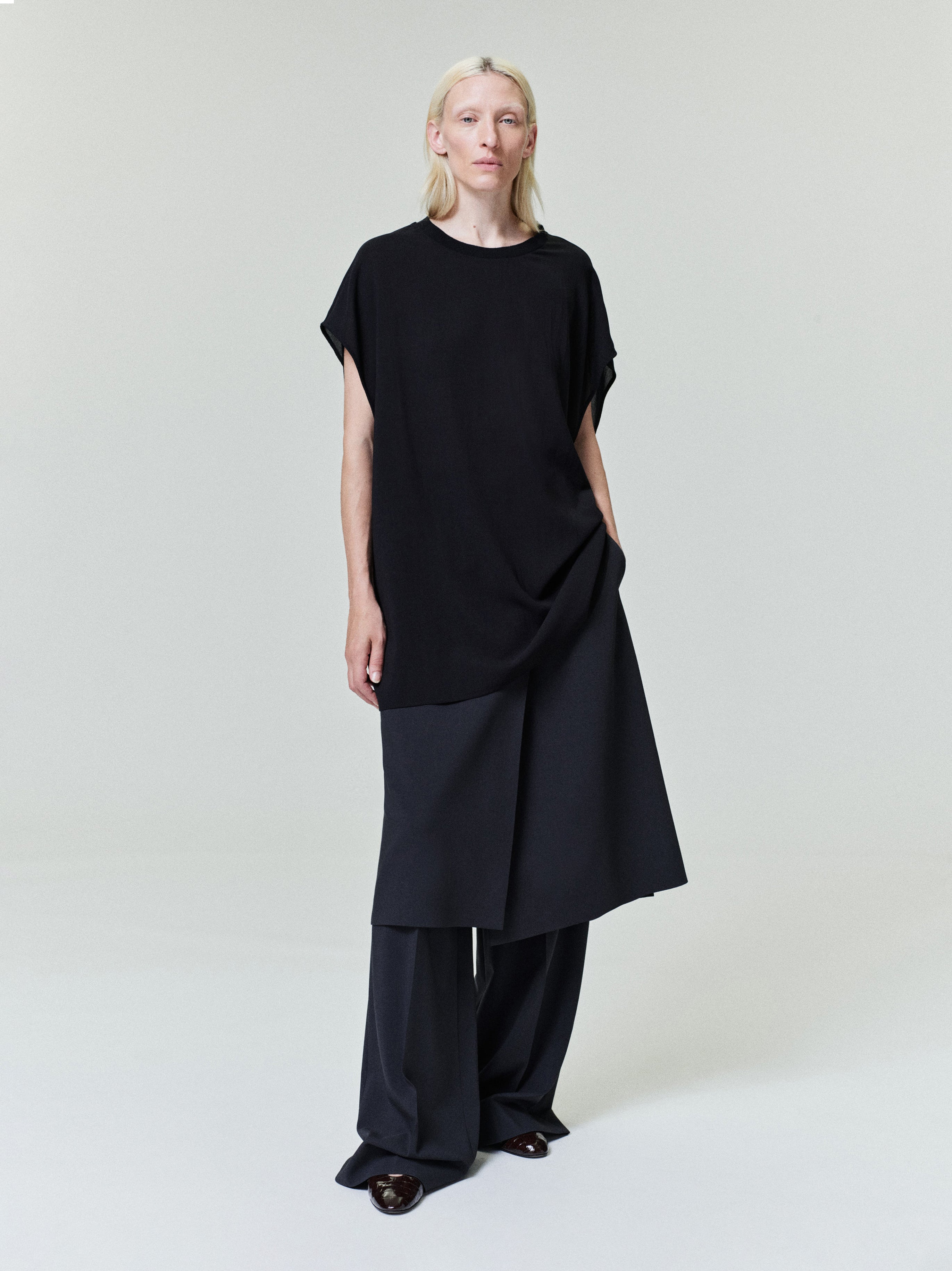 Person with pale blonde hair wearing loose black asymmetrical layered top and wide black pants on light gray background