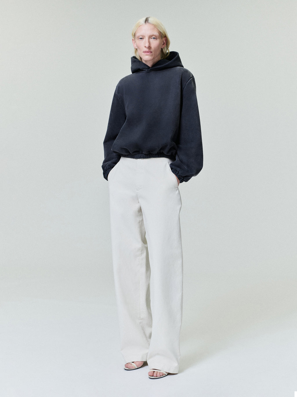 Person with light blonde hair wearing black oversized hoodie and wide white pants with open-toe heels on plain light background