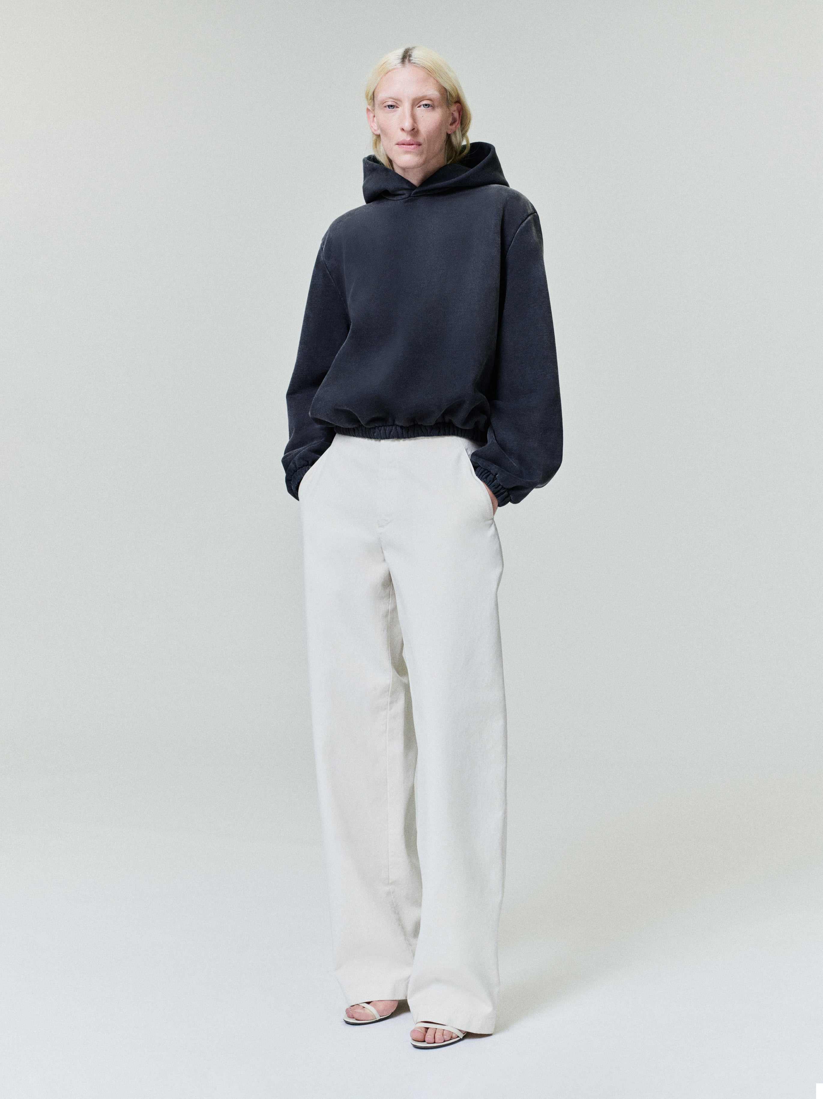 Person with light blonde hair wearing black oversized hoodie and wide white pants with open-toe heels on plain light background