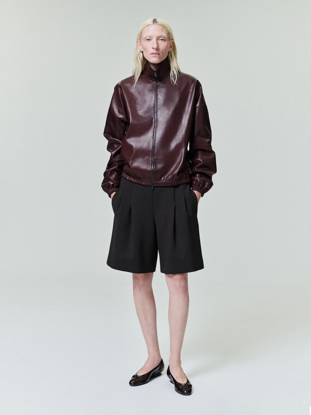 Person with straight pale blonde hair wearing dark brown zip-up leather jacket and loose black knee-length shorts on light background