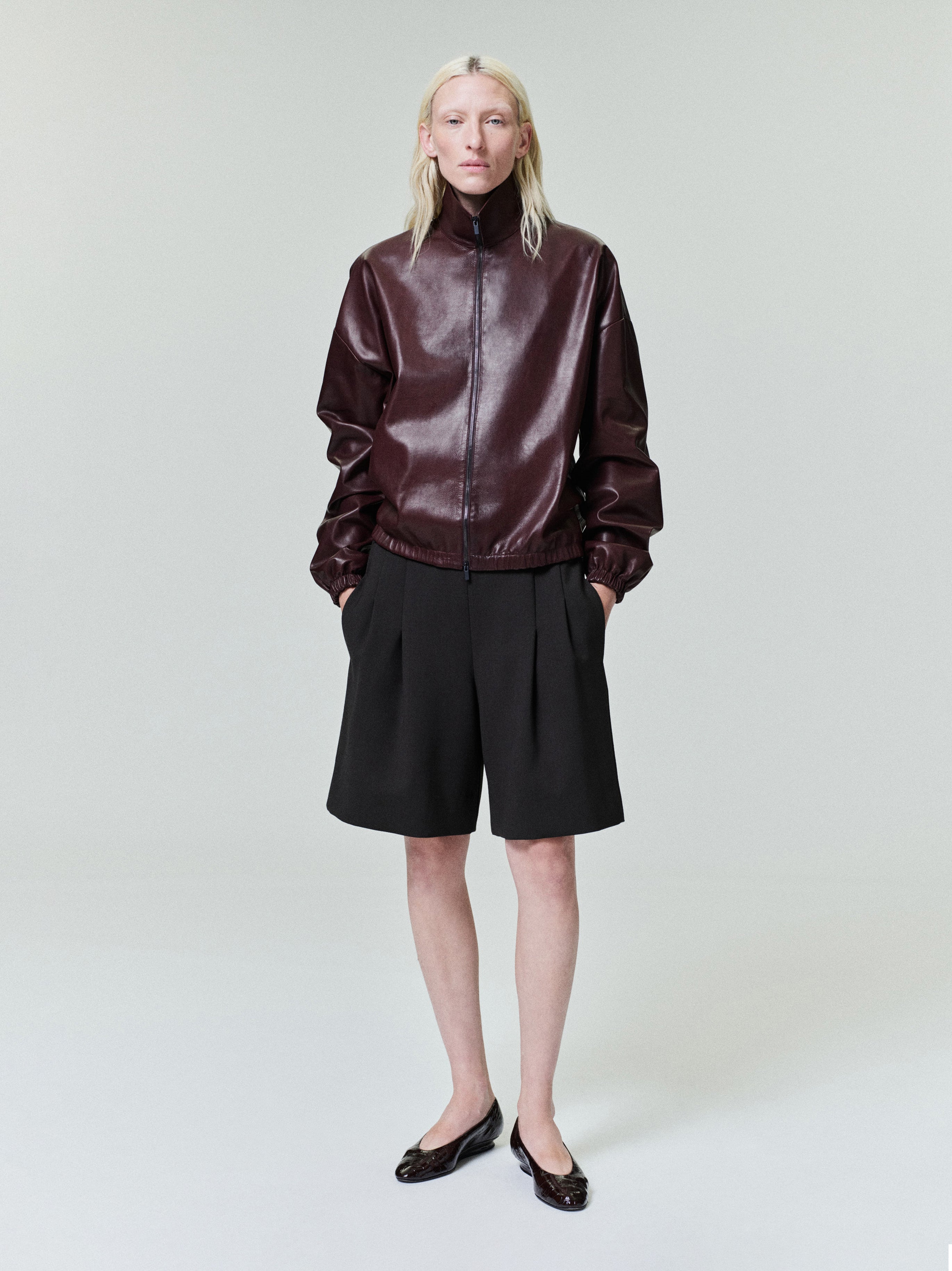 Person with straight pale blonde hair wearing dark brown zip-up leather jacket and loose black knee-length shorts on light background