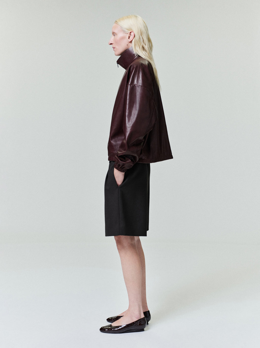 Person with straight pale blonde hair wearing dark burgundy leather jacket and black knee-length shorts on light gray background