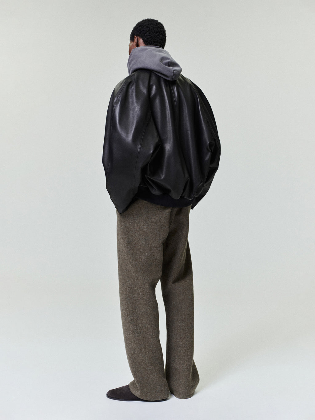 Man seen from behind wearing a loose black leather jacket over a gray hooded sweatshirt and wide brown trousers with dark shoes