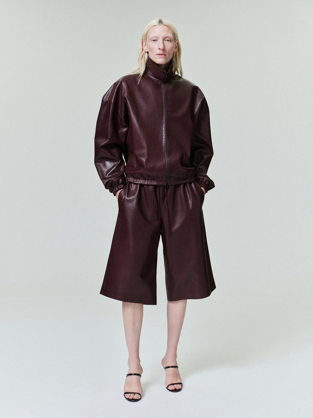 Person with pale blonde hair wearing dark burgundy leather zip-up jacket and matching wide knee-length shorts on light background