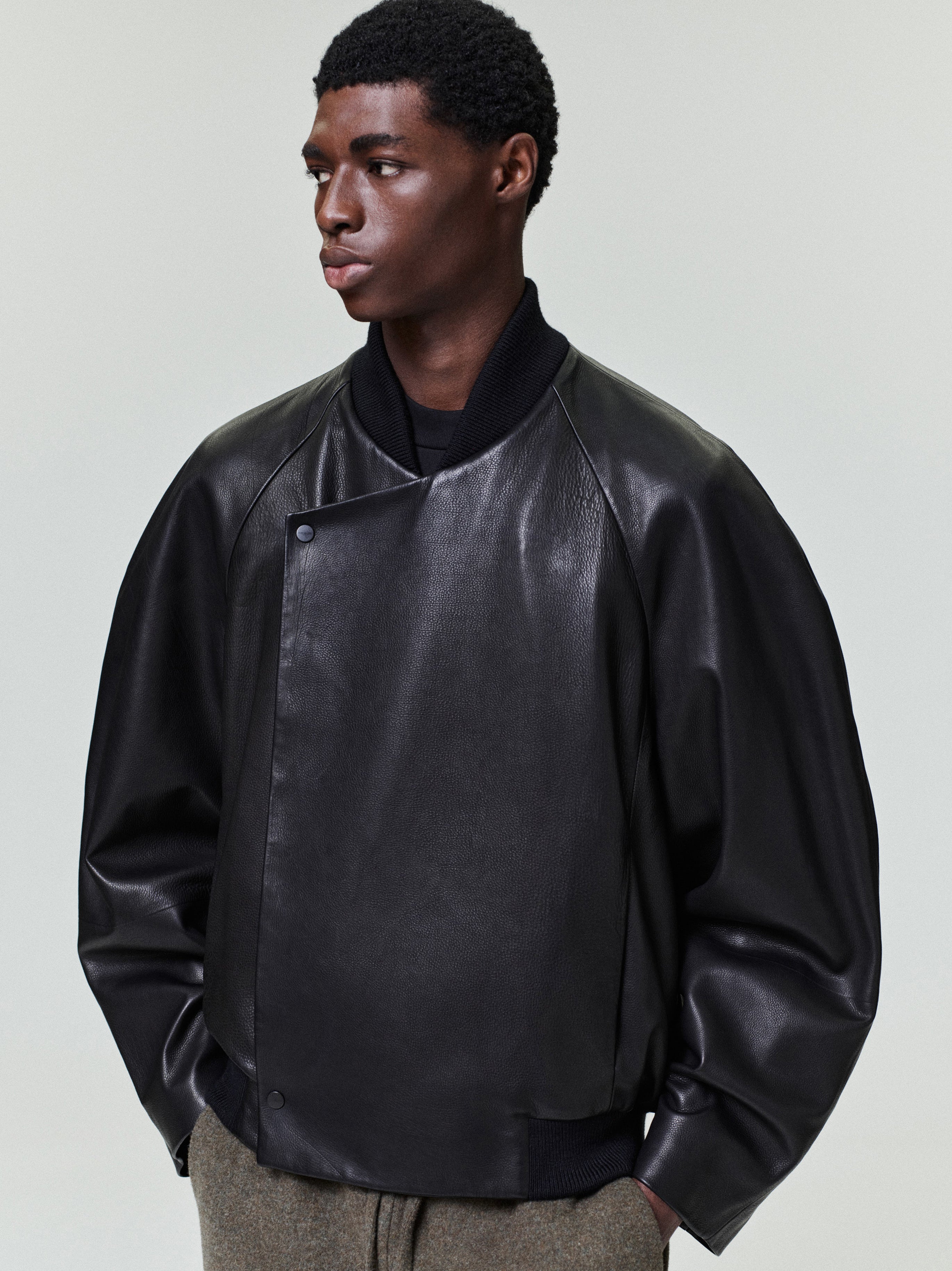 Man wearing a loose black leather bomber jacket with a high collar and ribbed cuffs over gray trousers