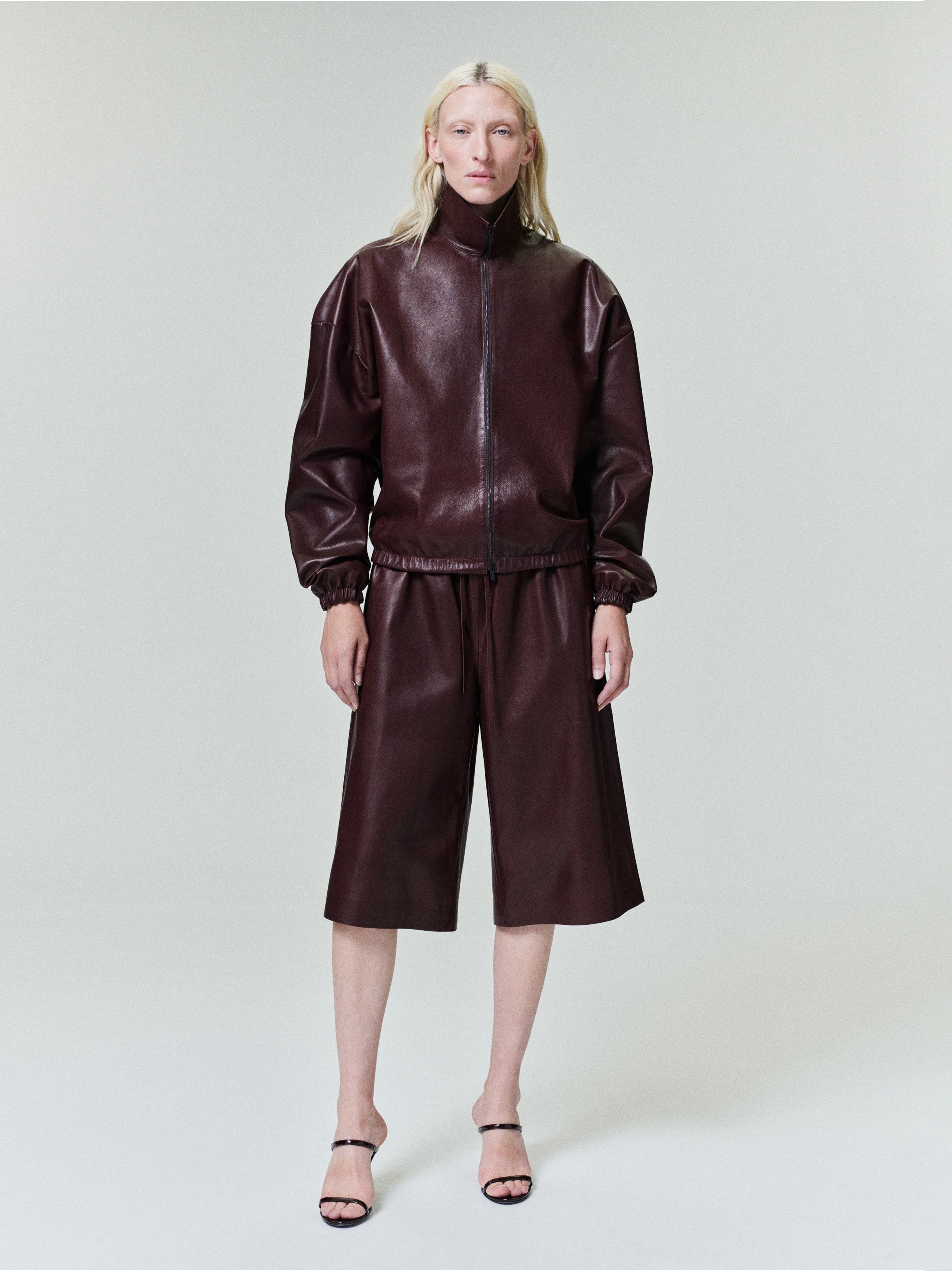 Person with straight pale blonde hair wearing dark brown leather zip-up jacket and matching wide knee-length shorts on pale background