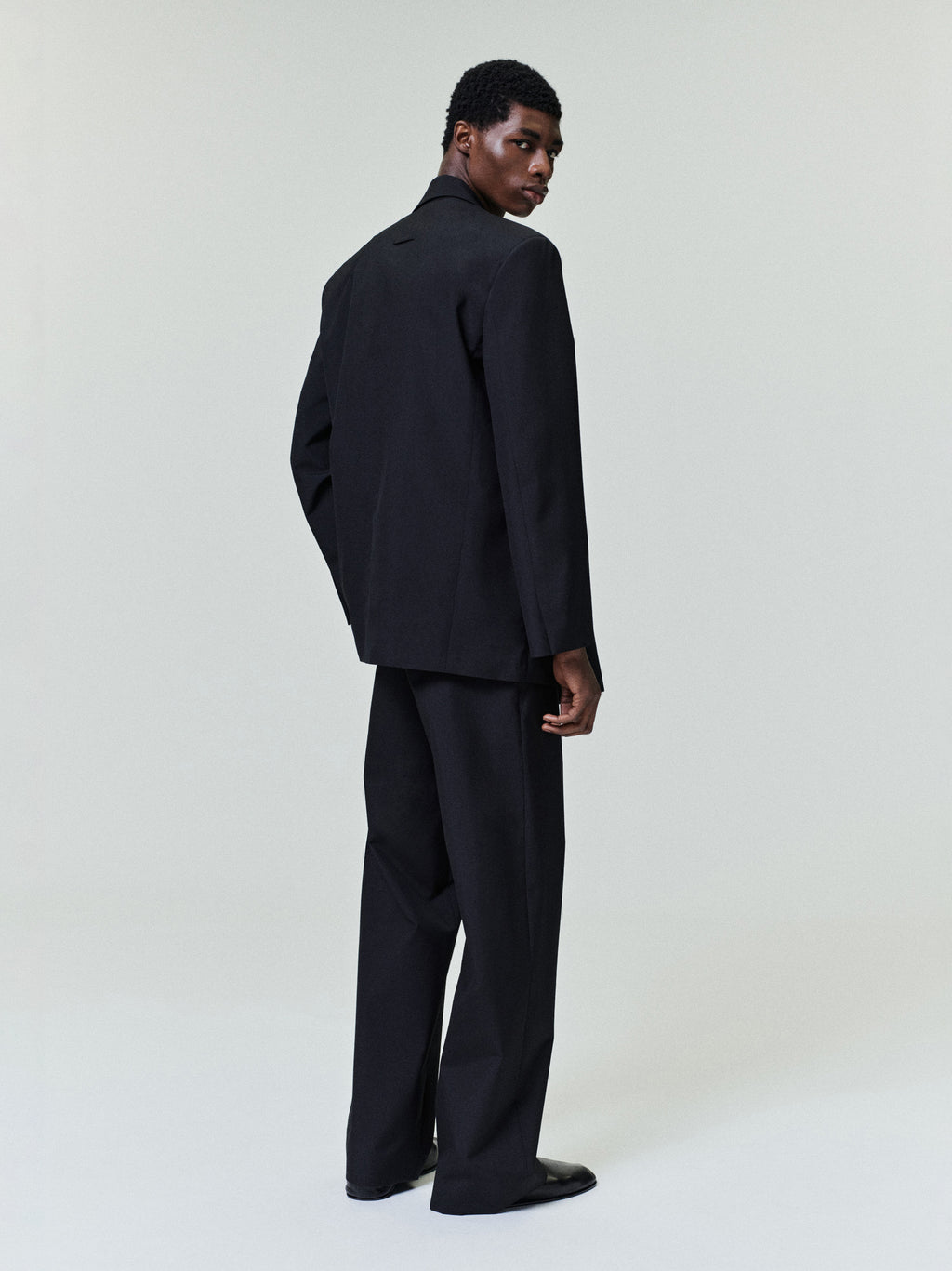 male model wearing black wool poplin casual flat front pants with relaxed fit and side seam pockets viewed from the back