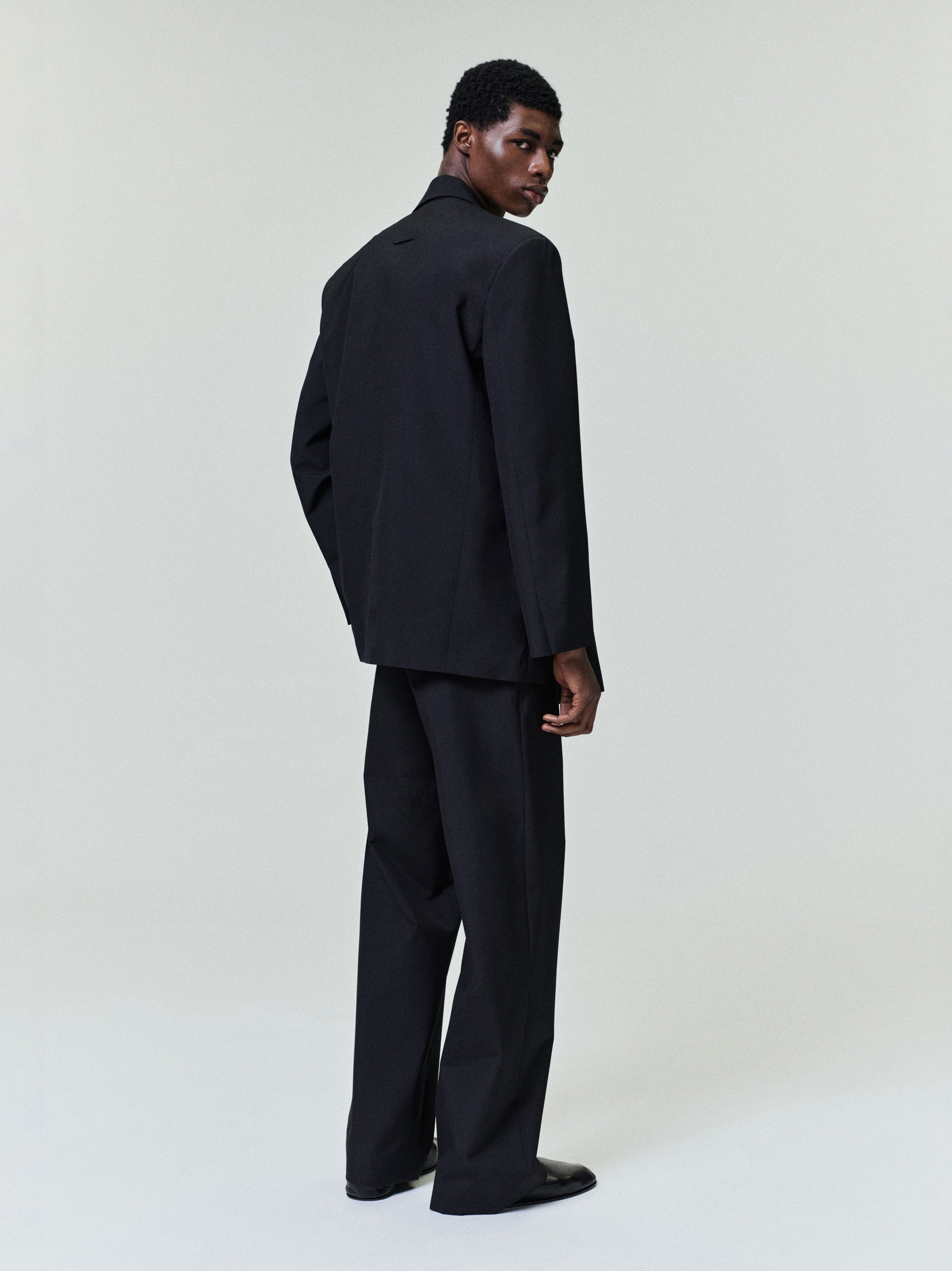 Man posing in a relaxed-fit black blazer and wide-leg black trousers viewed from behind against a plain background