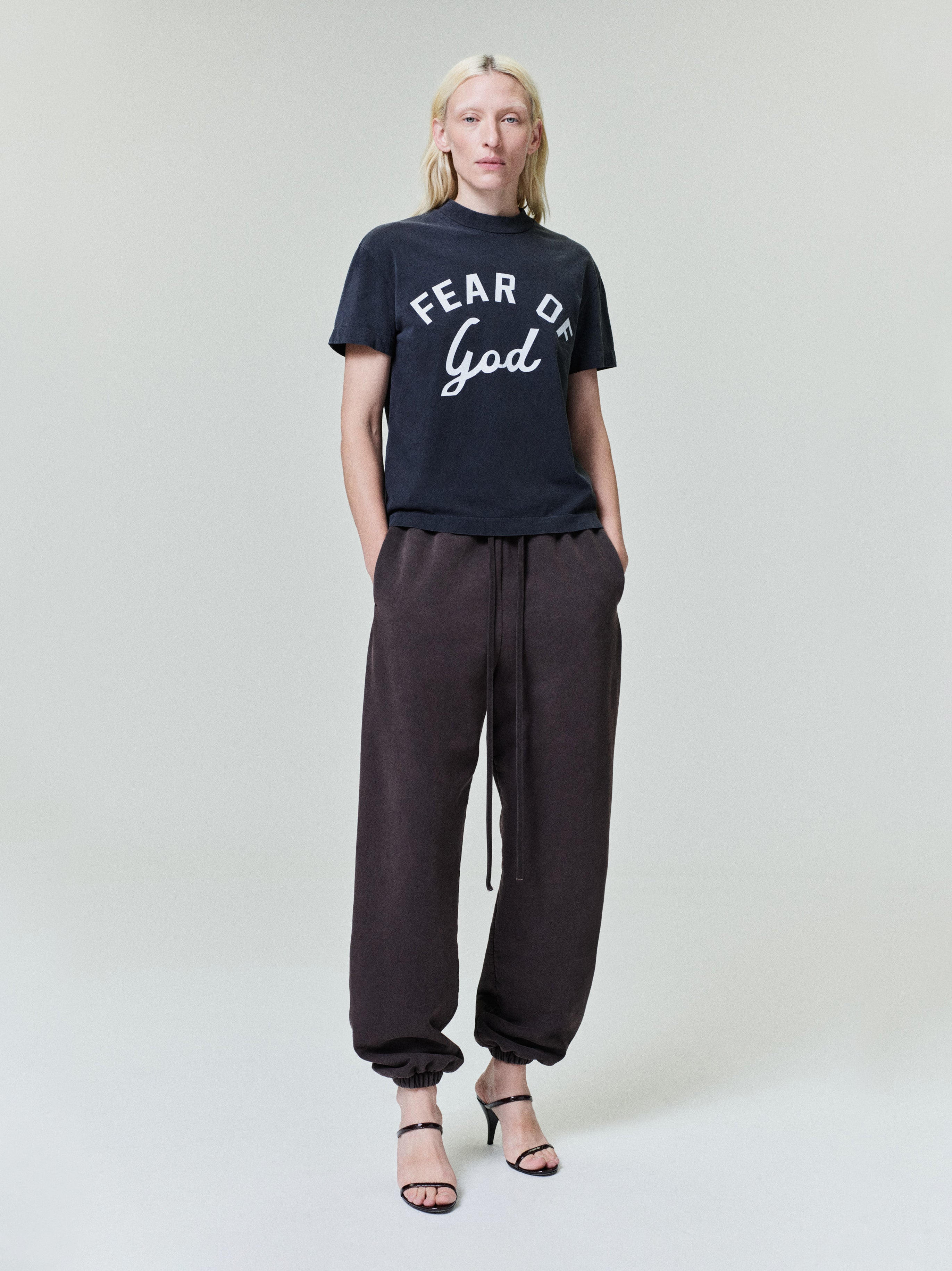 Person with light blonde hair wearing black "Fear of God" t-shirt, dark loose jogger pants, and black open-toe heels on gray background